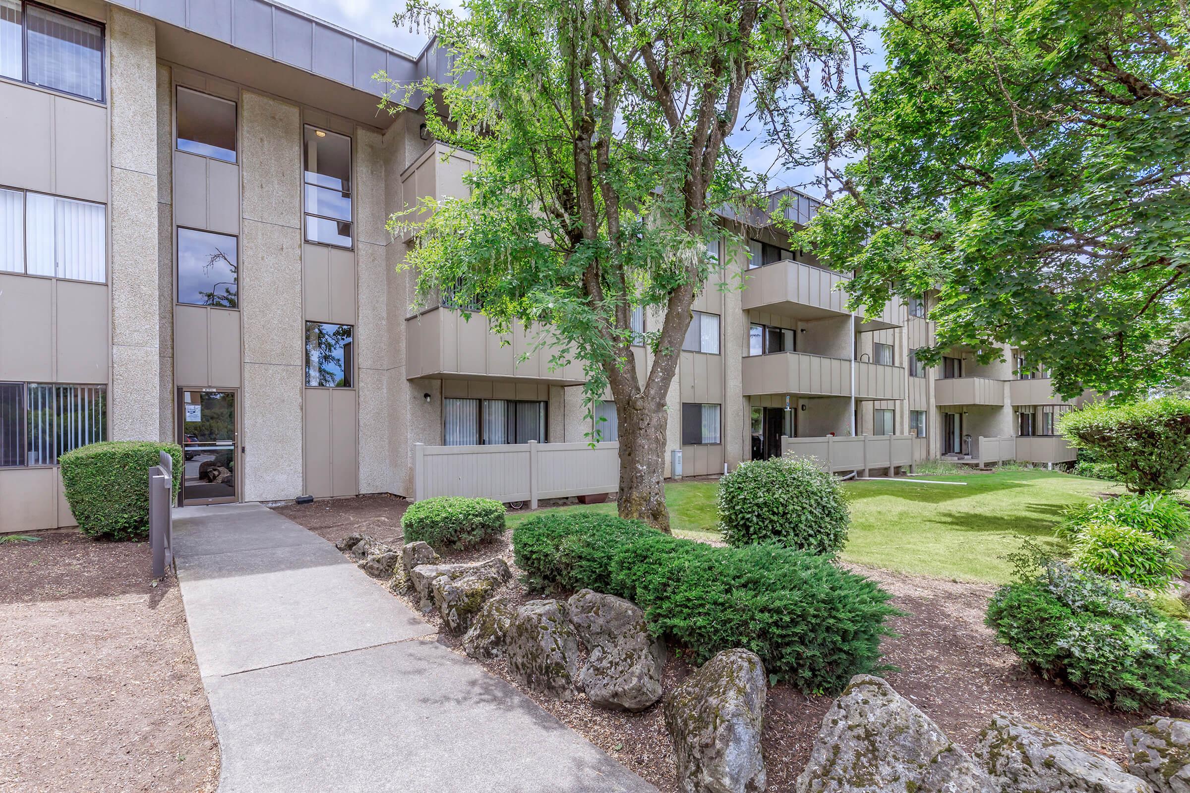 An exterior view of a multi-story apartment building surrounded by greenery. The building features a beige facade with balconies. A pathway leads from the entrance, flanked by bushes and landscaping. Trees provide shade, contributing to a pleasant, inviting environment.
