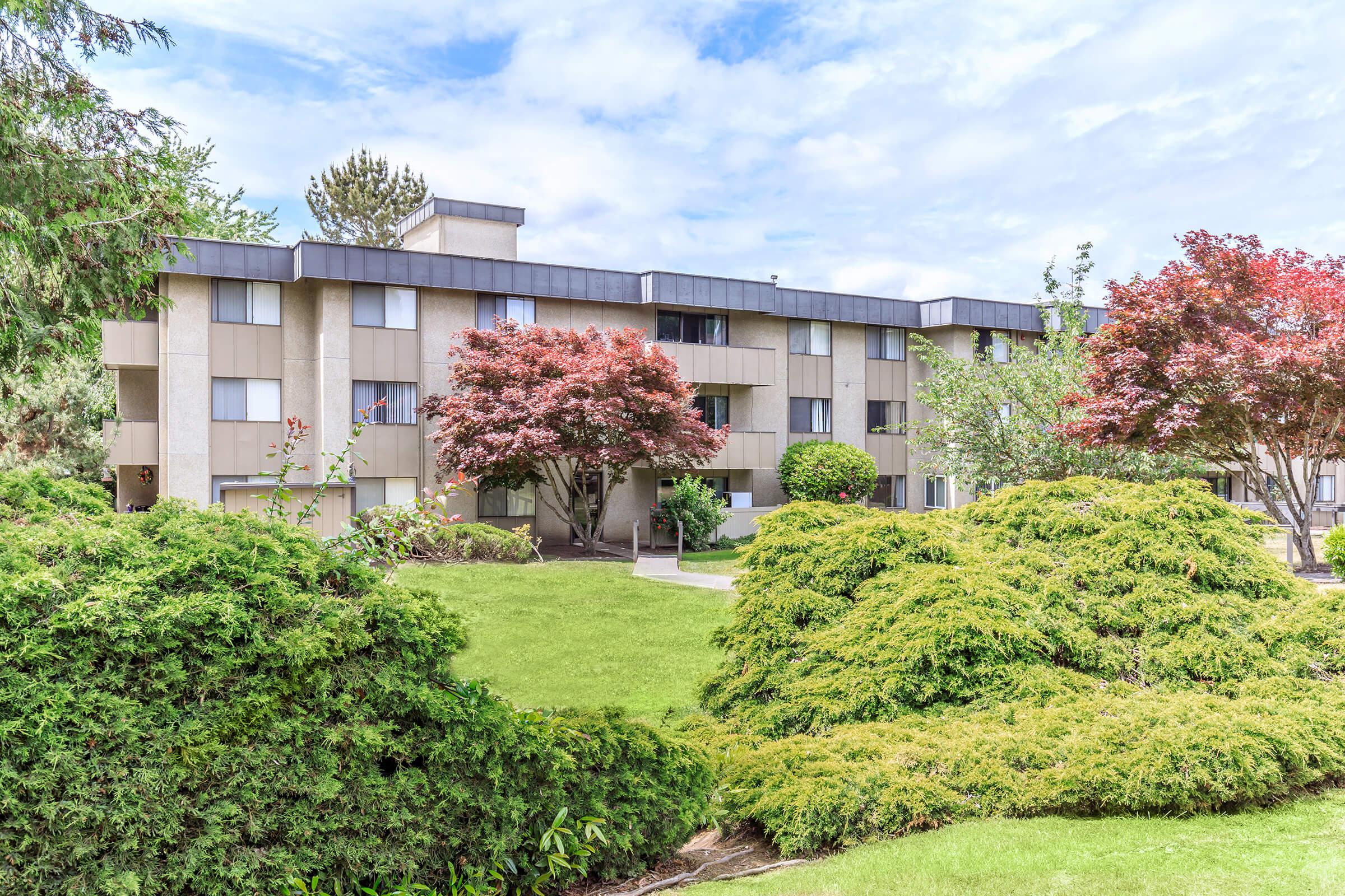 A multi-story apartment building surrounded by well-maintained green landscaping, including bushes and trees. The exterior features rectangular windows and a minimalist design, with vibrant red foliage from nearby trees adding color to the scene. The sky is partly cloudy.