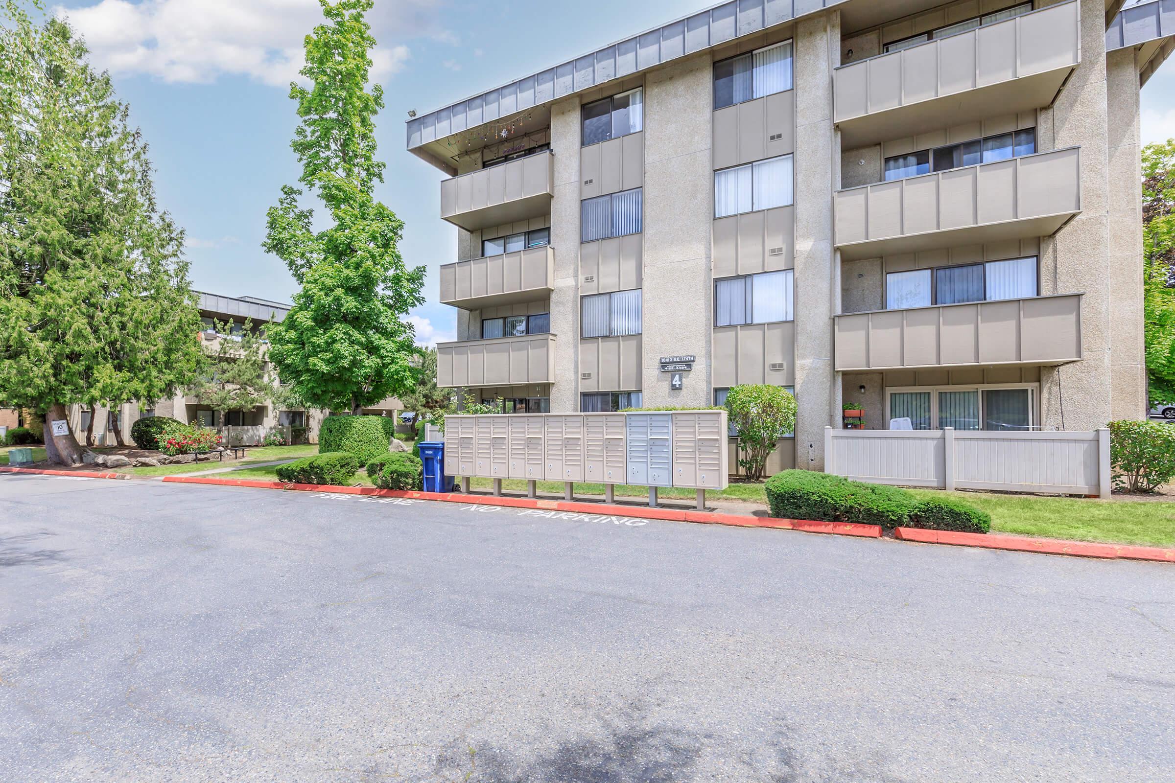 A view of a multi-story apartment building surrounded by trees and landscaped areas. The building features several balconies and windows, with a mailbox area in the foreground. The pavement in front indicates parking spaces. The sky is partly cloudy, adding to a calm residential atmosphere.