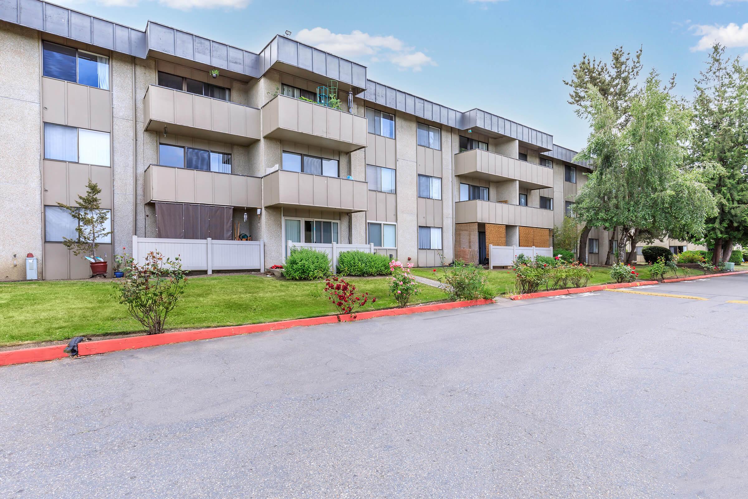 A multi-story apartment building with a modern architectural design, featuring balconies and landscaped greenery in front. The facade has a mix of light and dark tones, with well-maintained flower beds lining the walkways. The surrounding area includes paved paths and trees under a clear sky.