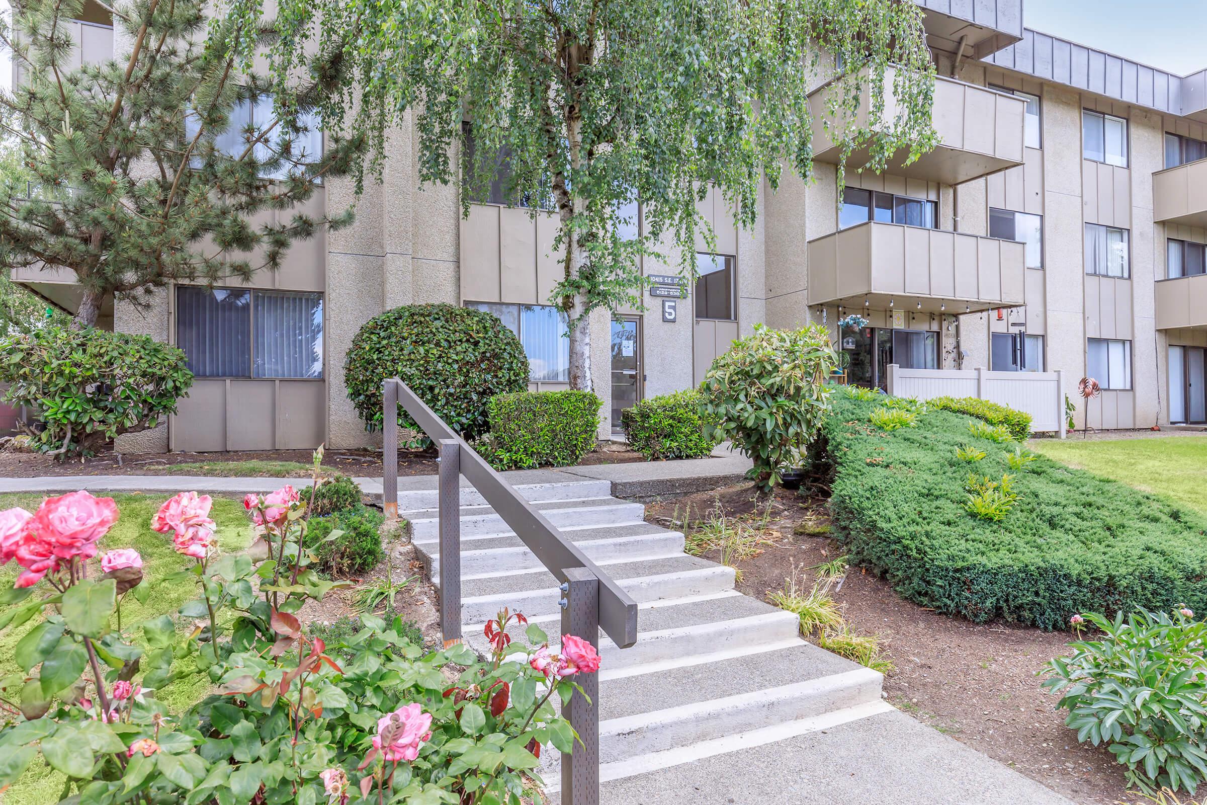 A landscaped entryway with a set of stairs leading up to a multi-story building. Lush greenery, including bushes and a tree, surrounds the area. Pink roses are blooming in the foreground, adding color to the scene. The building features large windows and a modern architectural style.