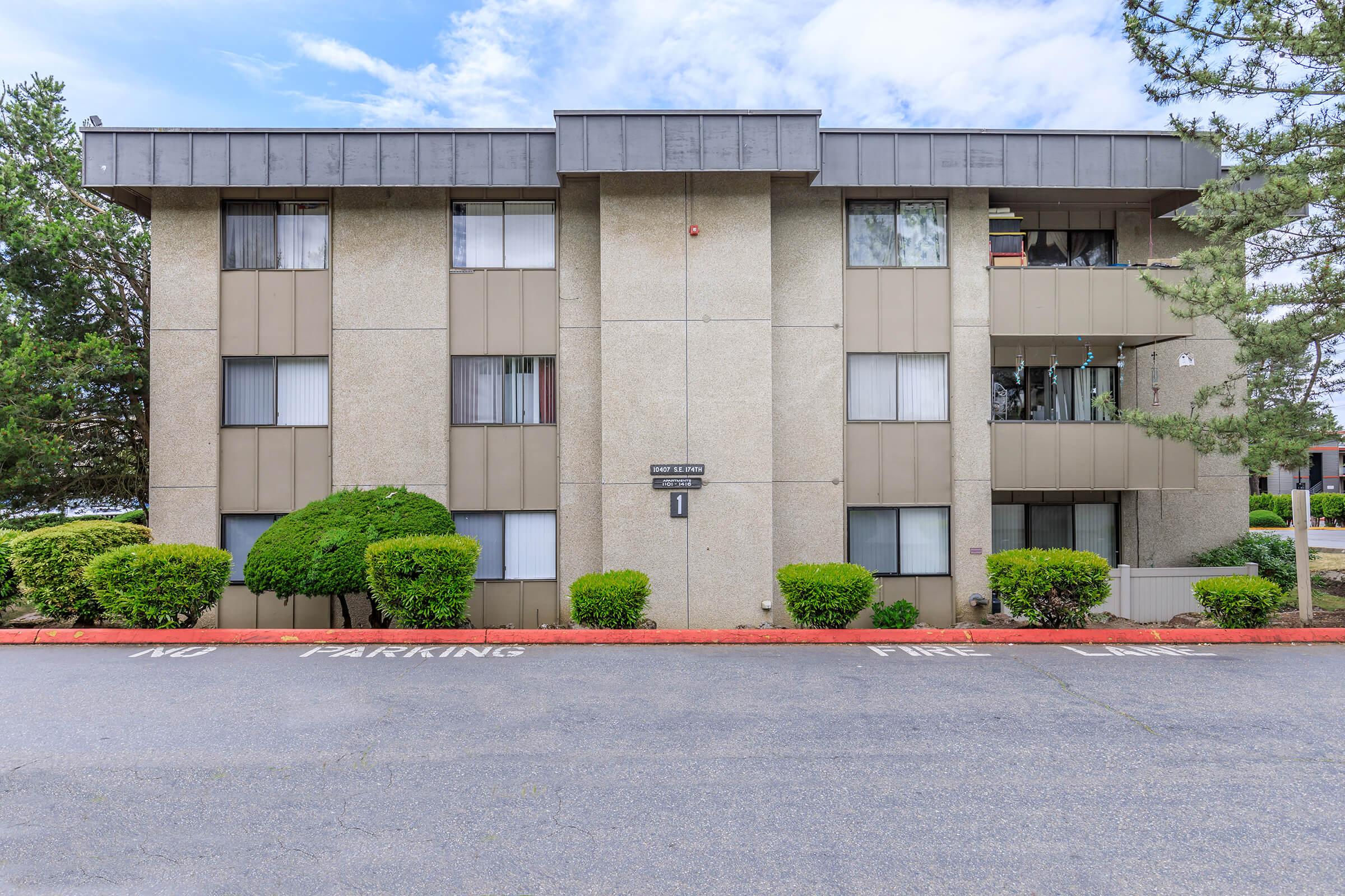 A three-story apartment building with a beige exterior and dark gray accents. The front features multiple windows with white curtains. A small sign displays the building number, and there are neatly trimmed bushes along the sidewalk. A parking area is visible in front with markings indicating no parking in certain spots.