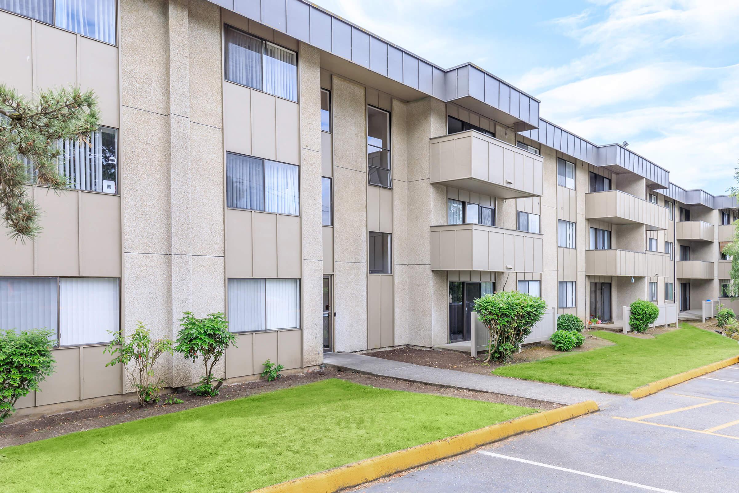 A modern, multi-story residential building with beige concrete exteriors and multiple balconies. Lush greenery surrounds the entrance, and a driveway runs alongside the building. The sky is clear and blue, adding to the pleasant ambiance of the scene.