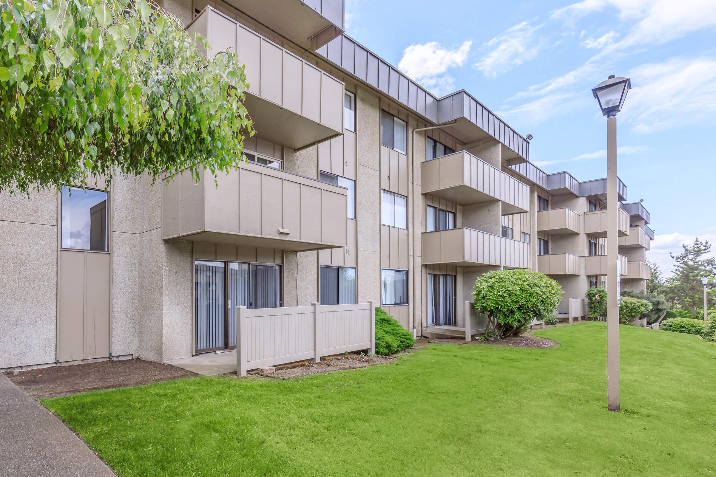 A modern apartment complex with multiple levels, featuring balconies and large windows. The building is surrounded by well-kept green lawns and shrubs. A pathway runs alongside the structure, with a street lamp visible. The sky is partly cloudy, indicating a bright day.