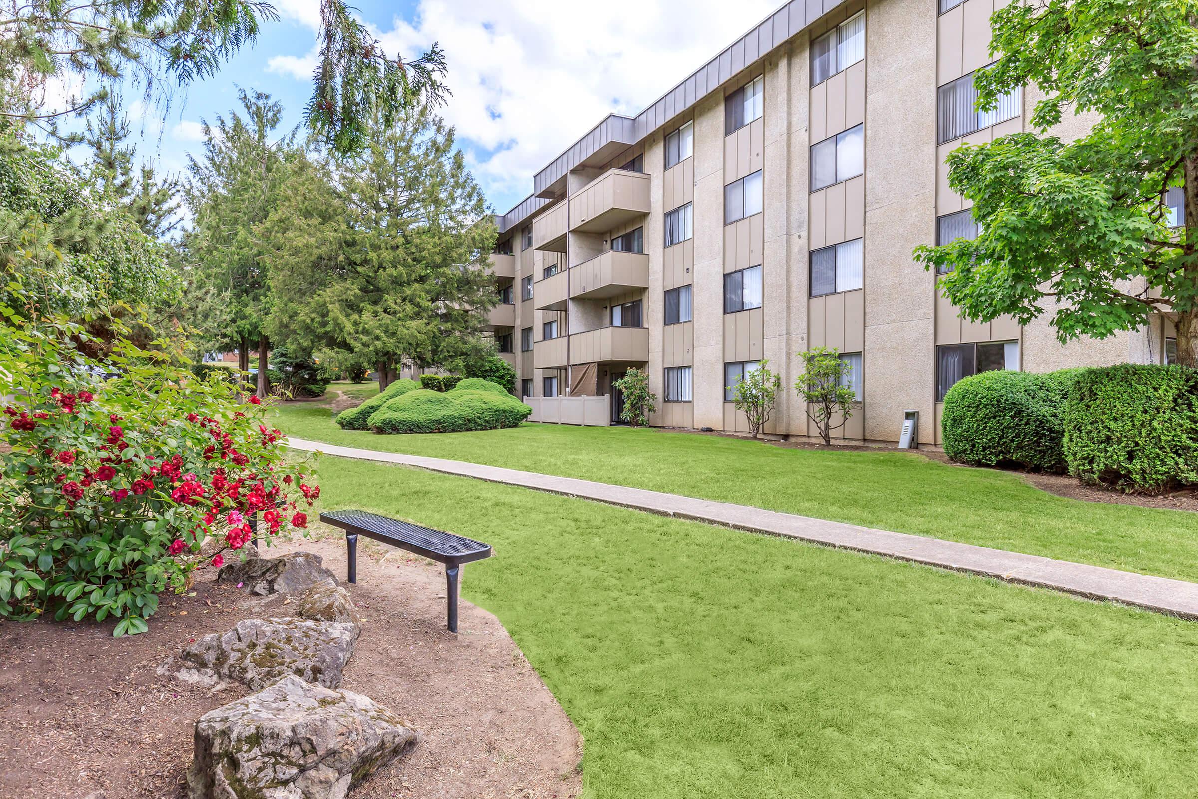 A well-maintained green lawn surrounds a multi-story apartment building. There are neatly trimmed bushes, rose bushes with vibrant red flowers, and a pathway leading to the building. A bench is positioned in front of the flowers, creating a pleasant outdoor setting. Trees are visible in the background under a partly cloudy sky.