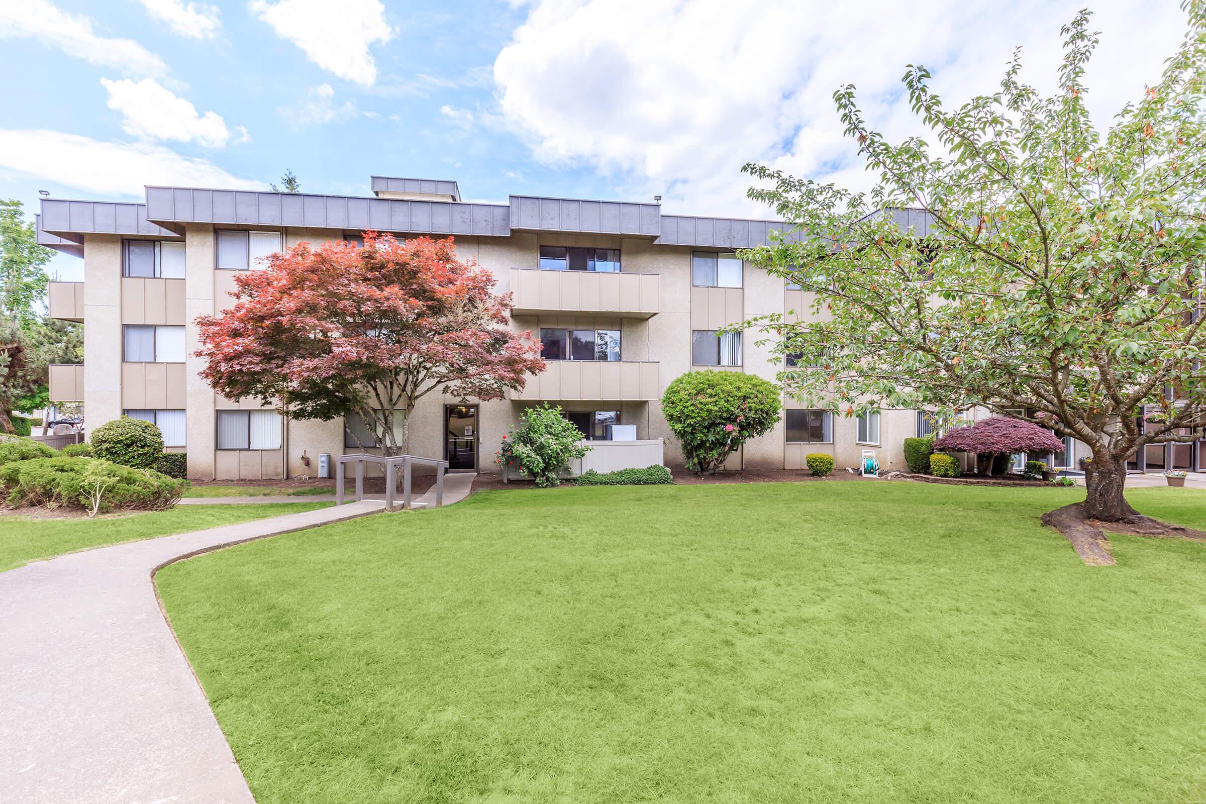 A multi-story residential building surrounded by well-maintained lawns and trees, including a red maple and a leafy green shrub. The sky is partly cloudy, and a paved pathway leads towards the entrance of the building. A peaceful outdoor setting is visible.