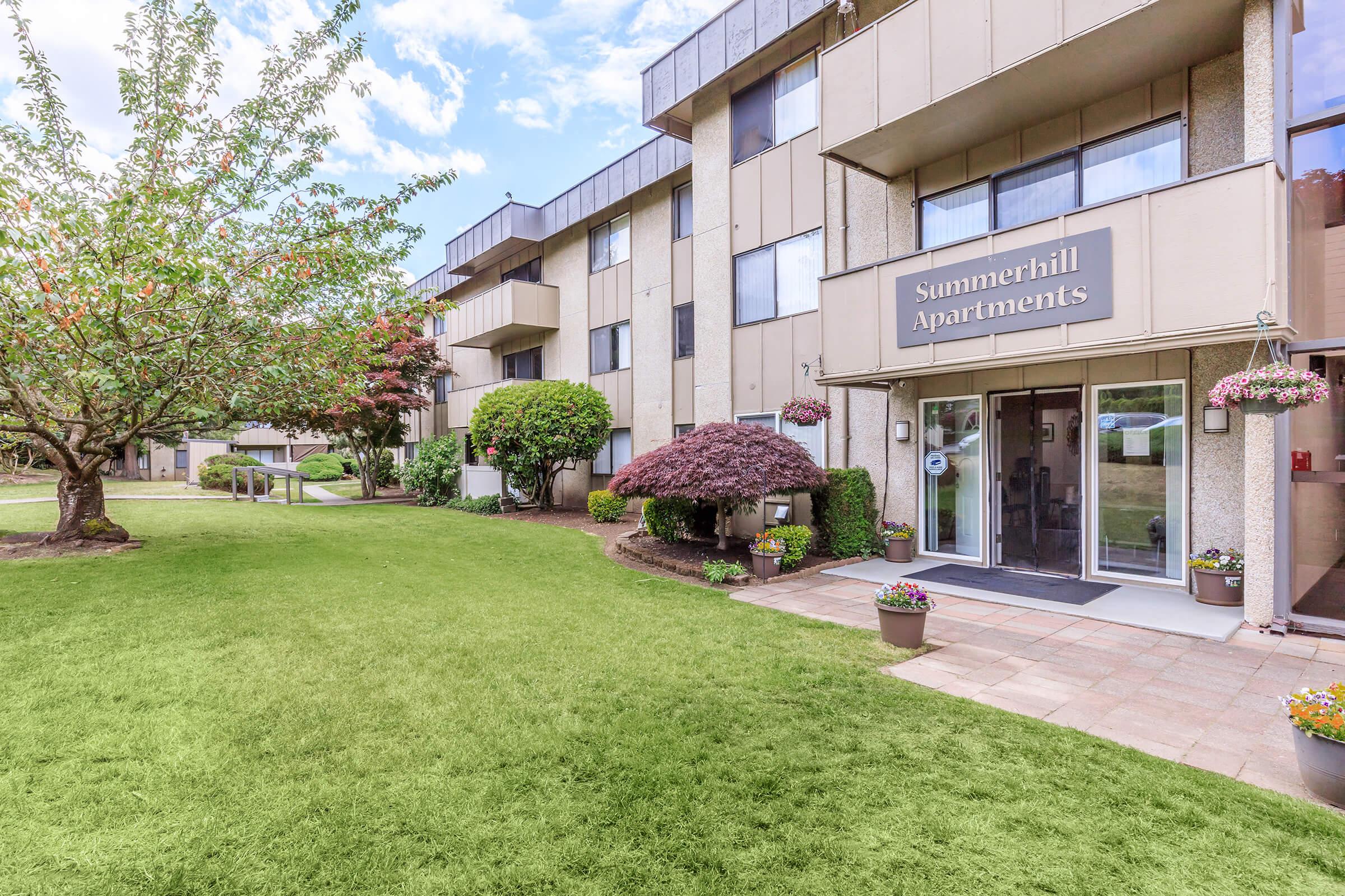 Front view of Summerhill Apartments featuring a well-maintained entrance with large glass doors, surrounded by lush greenery and colorful flower pots. A manicured lawn and trees are visible in the foreground, creating a welcoming atmosphere. The building is modern with balconies and a light-colored exterior.