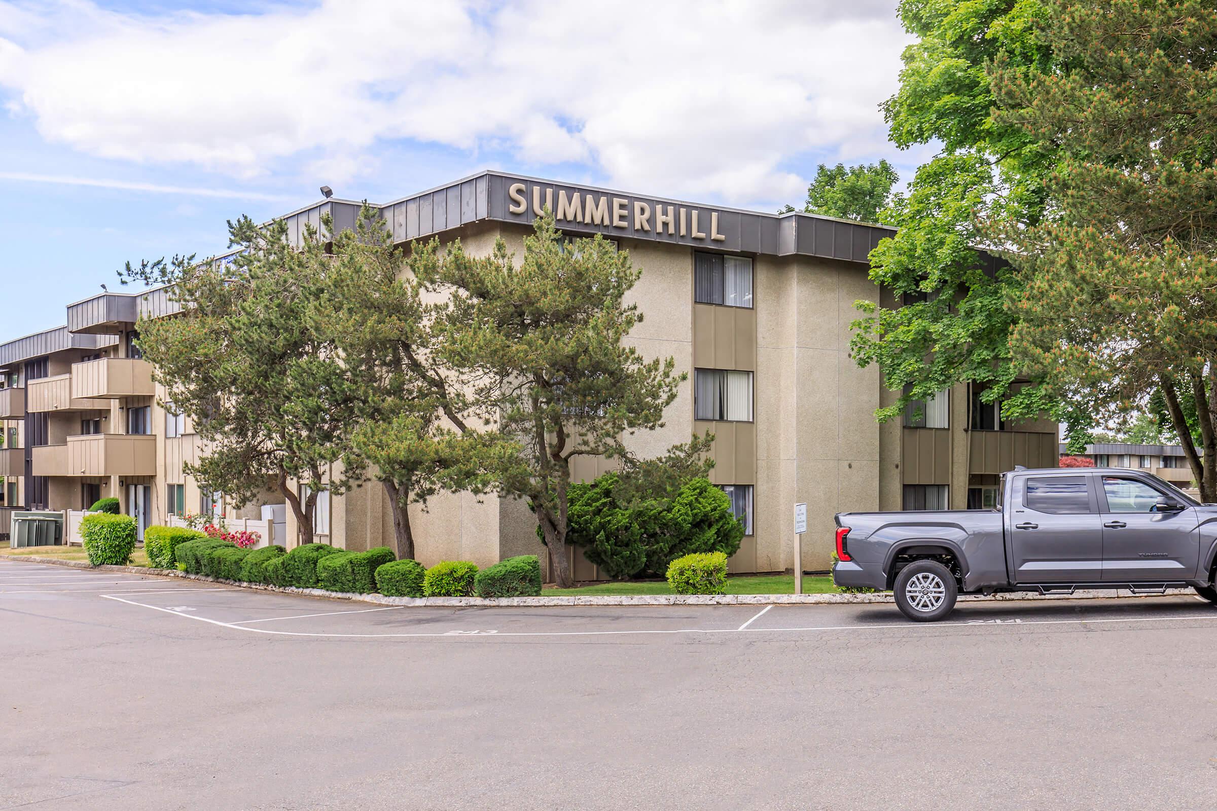 A three-story building with the name "SUMMERHILL" displayed on the front. The building has a mix of windows and beige siding, surrounded by well-maintained greenery. A gray pickup truck is parked in front, and the sky is partly cloudy.