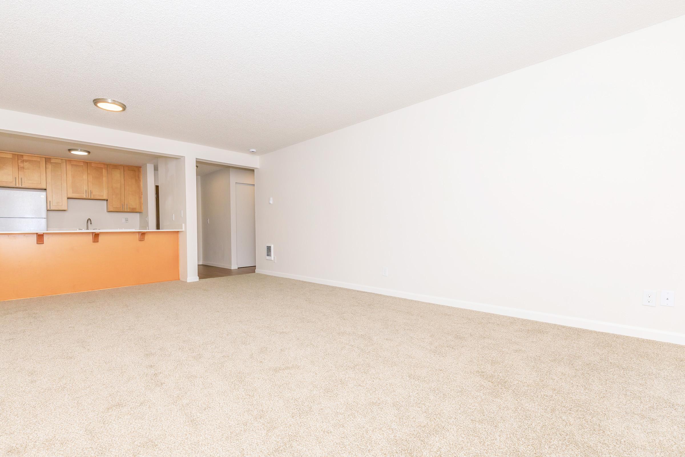 A spacious, empty living area with beige carpet and light-colored walls. To the left, a kitchenette is visible, featuring wooden cabinetry and a small bar area. A doorway on the right leads to another room or hallway, providing a minimalist and open atmosphere.