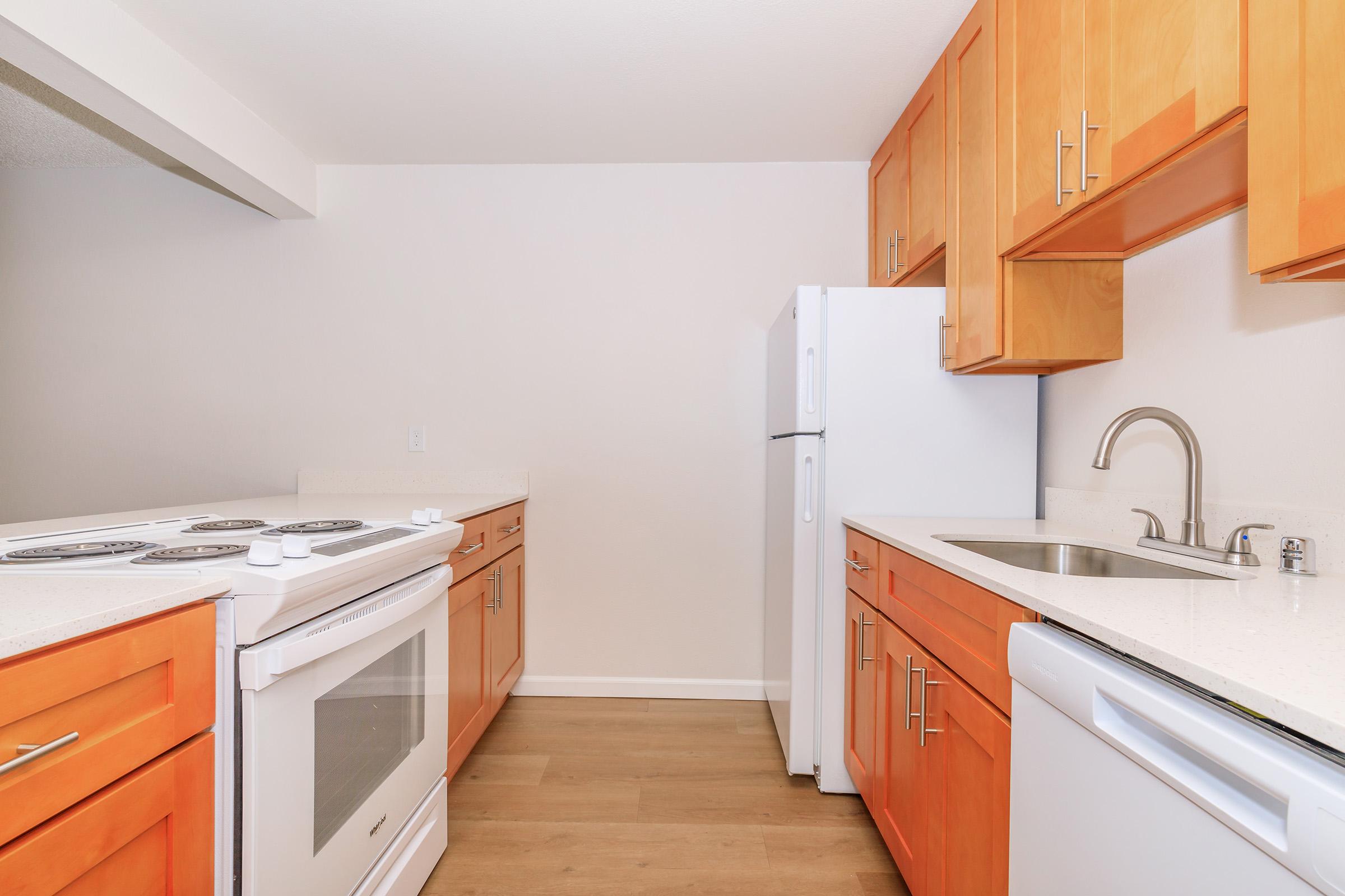 A modern kitchen with light-colored cabinets, a white refrigerator, an electric stove with an oven, and a sink. The countertops are light, and the flooring is a warm wood tone. The kitchen is well-lit and features a minimalistic design.
