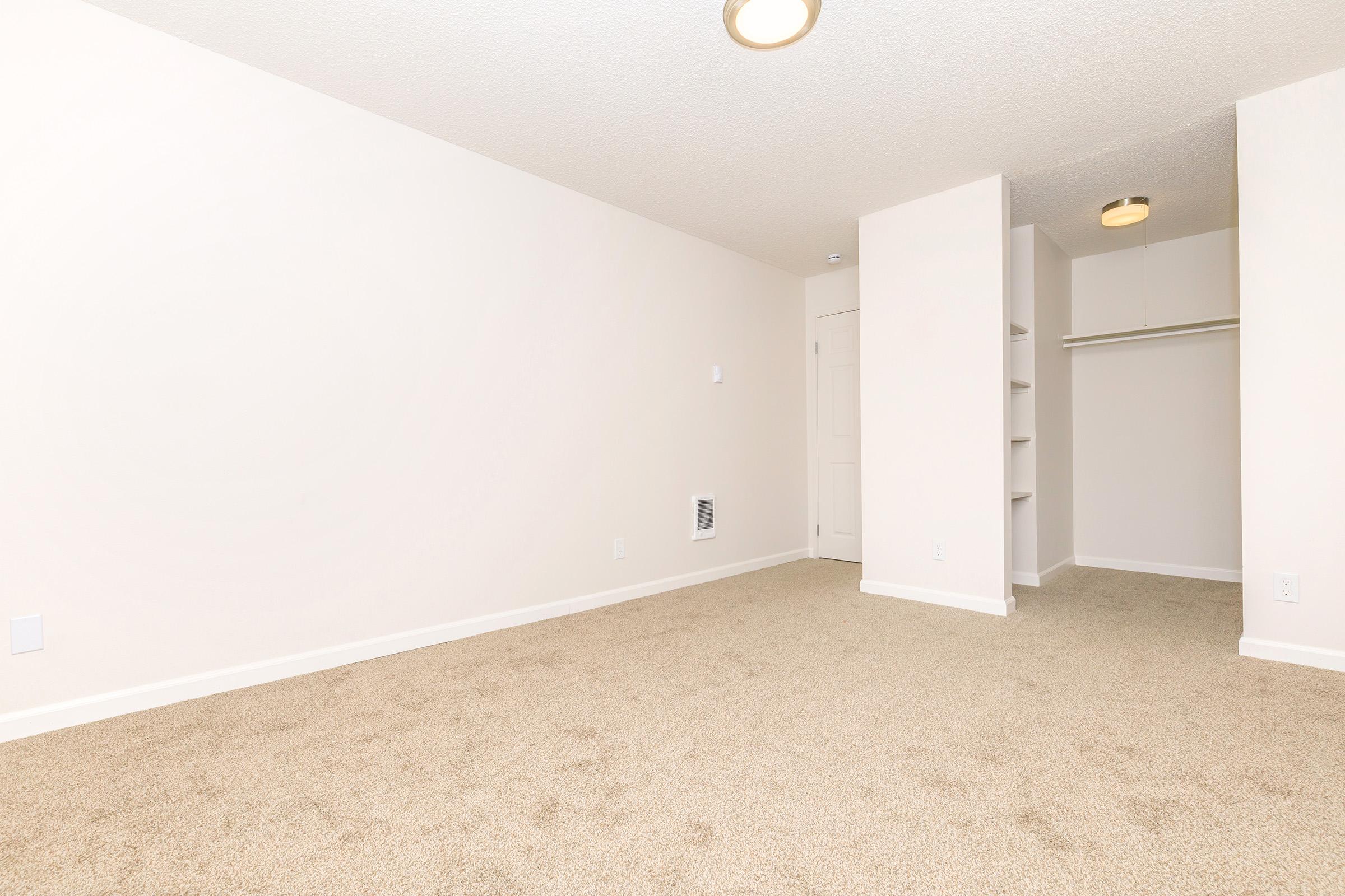 A spacious, empty room with beige carpet and light-colored walls. There is a closet with sliding doors on one side and a simple ceiling light fixture. The space is well-lit and ready for personal decoration or furniture arrangement.