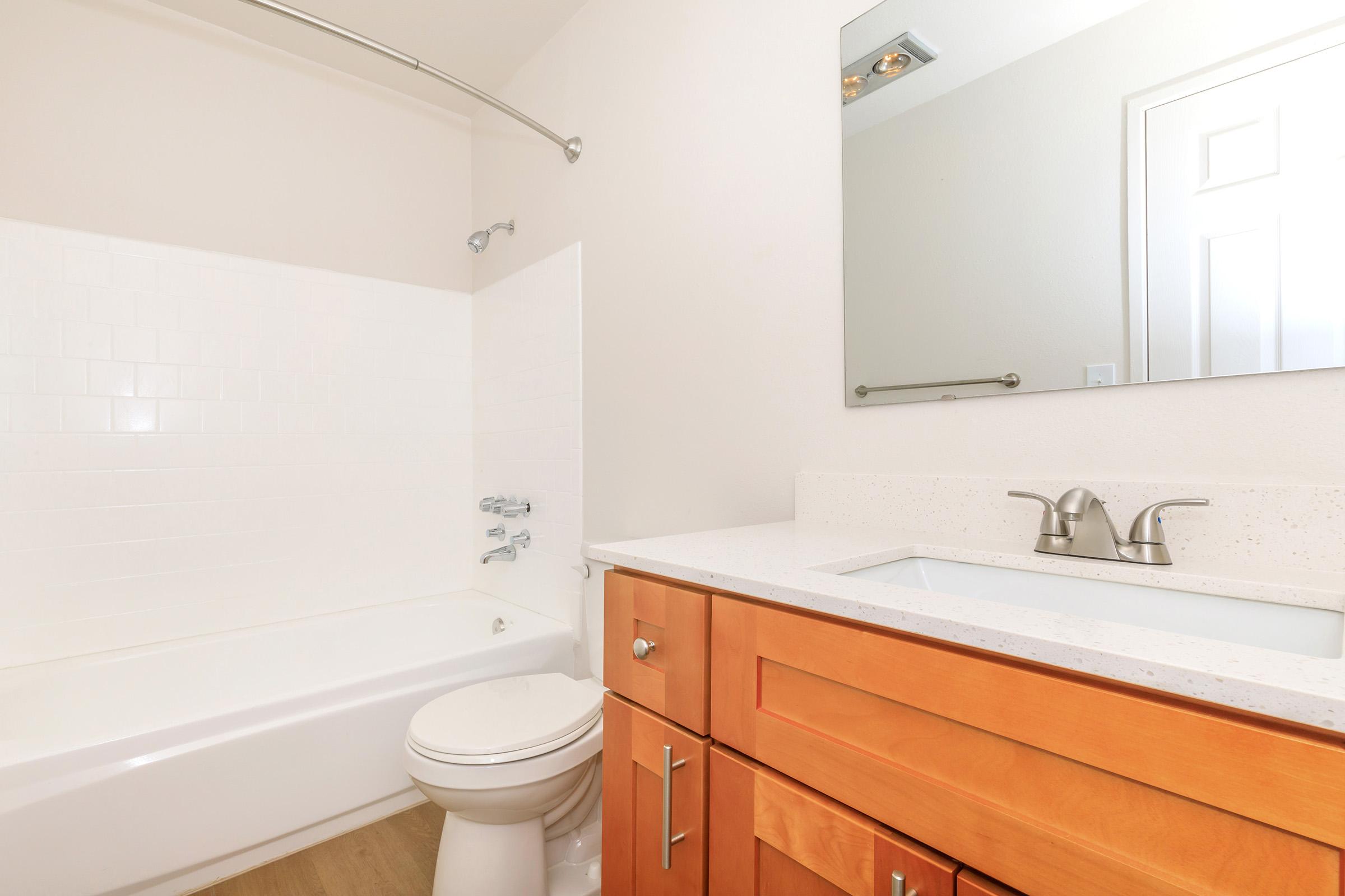 A clean and modern bathroom featuring a white bathtub with a shower, a white wall, a large mirror above a stylish sink, and wooden cabinetry below. The floor has a light wood finish, and the overall color scheme is bright and neutral.