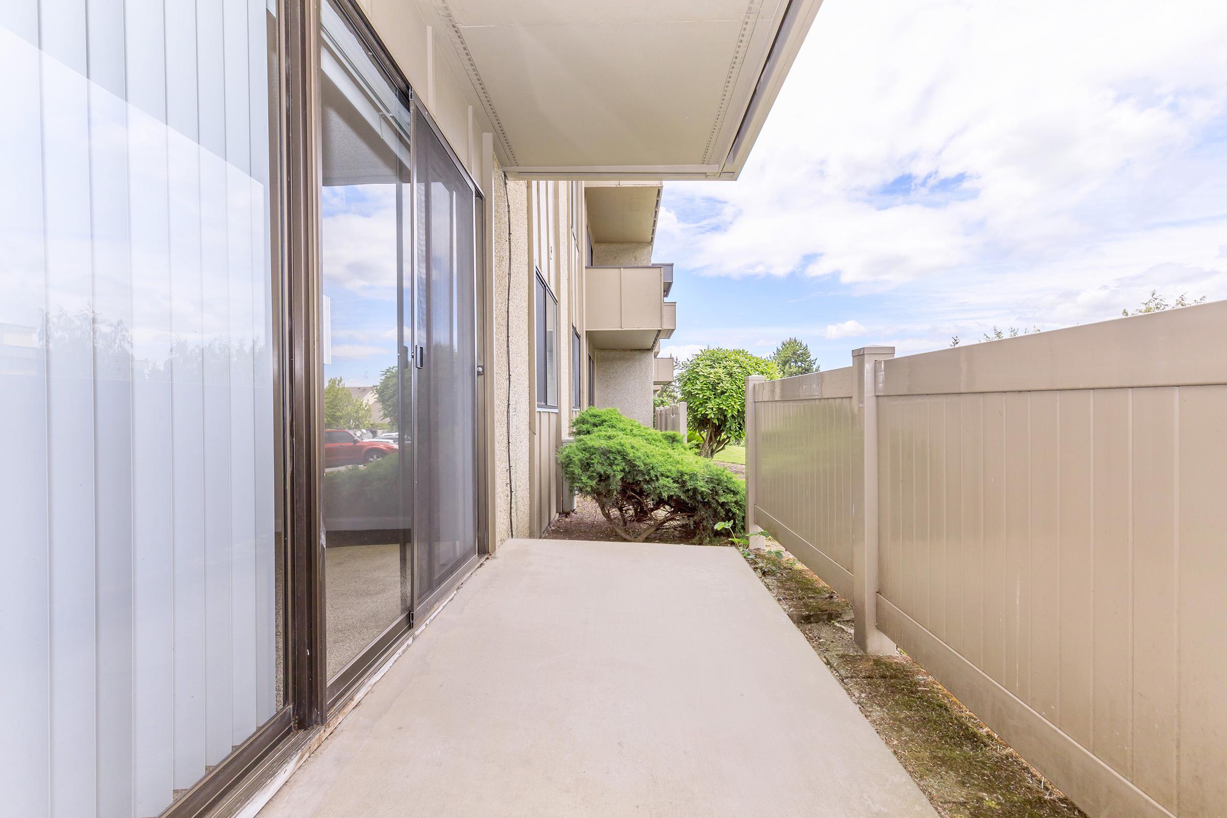 A view of a walkway leading to a building, featuring a smooth concrete path bordered by a low fence and greenery. The scene is illuminated by natural light, with a partly cloudy sky visible in the background. The setting conveys a quiet and peaceful atmosphere.