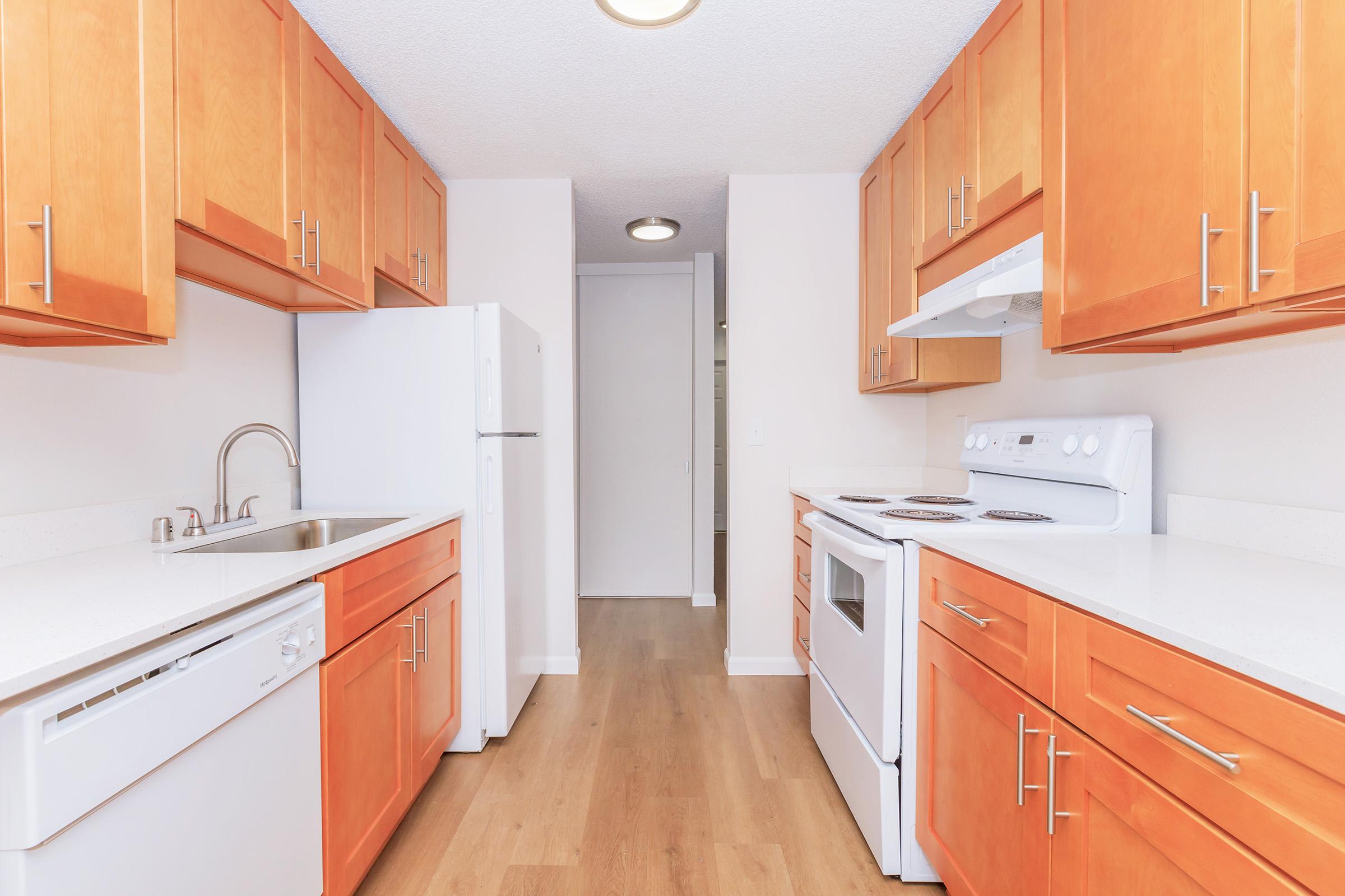 Bright and modern kitchen featuring wooden cabinets, white appliances including a refrigerator, stove, and dishwasher, and a spacious countertop. The area has wooden flooring and neutral-colored walls, creating an open and inviting atmosphere. A door leads to another room at the end of the hallway.