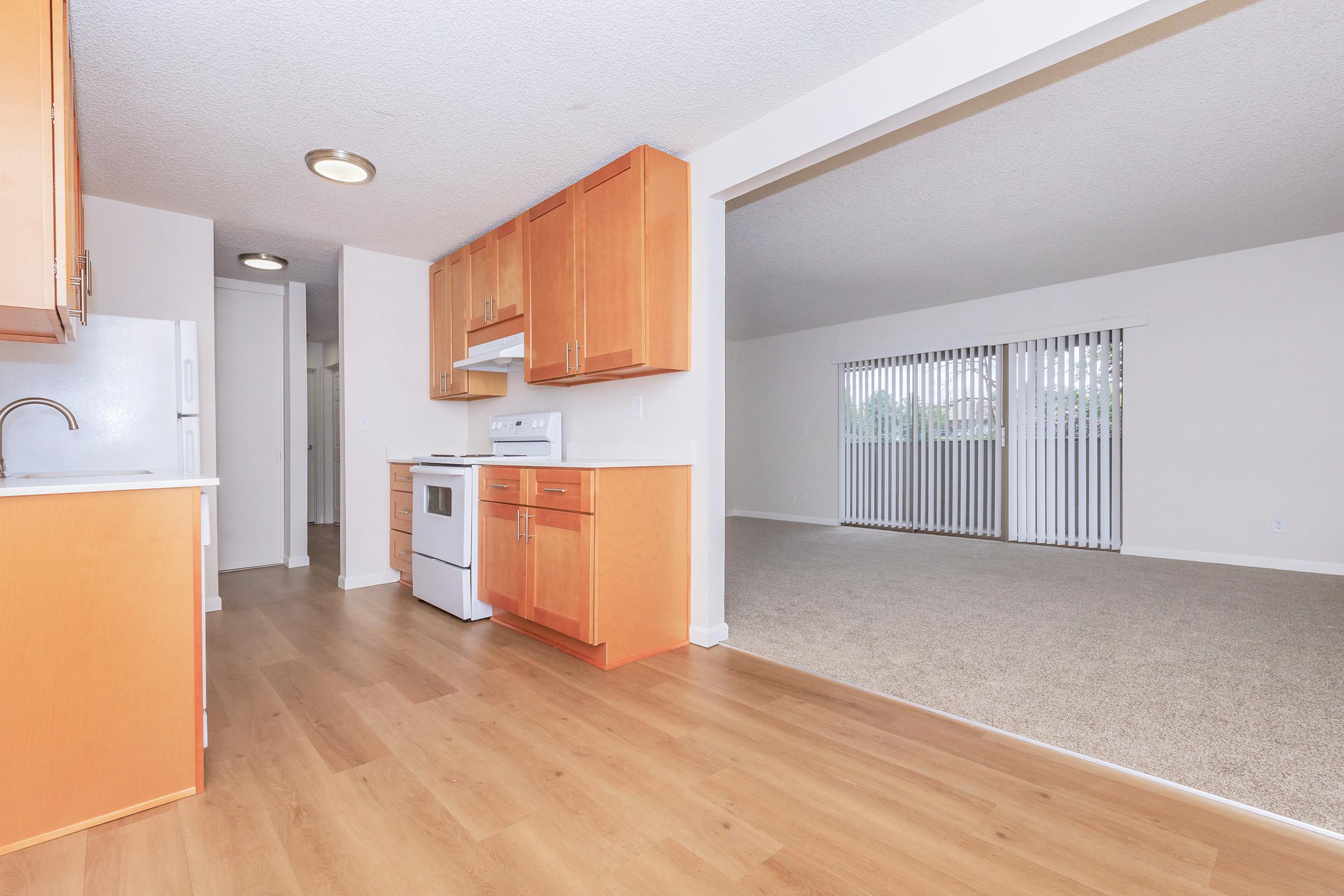 A spacious kitchen featuring wooden cabinets, a white stove, and a light-colored countertop. The room opens up to a living area with beige carpet and large windows covered by vertical blinds, allowing ample natural light. The overall ambiance is bright and inviting.