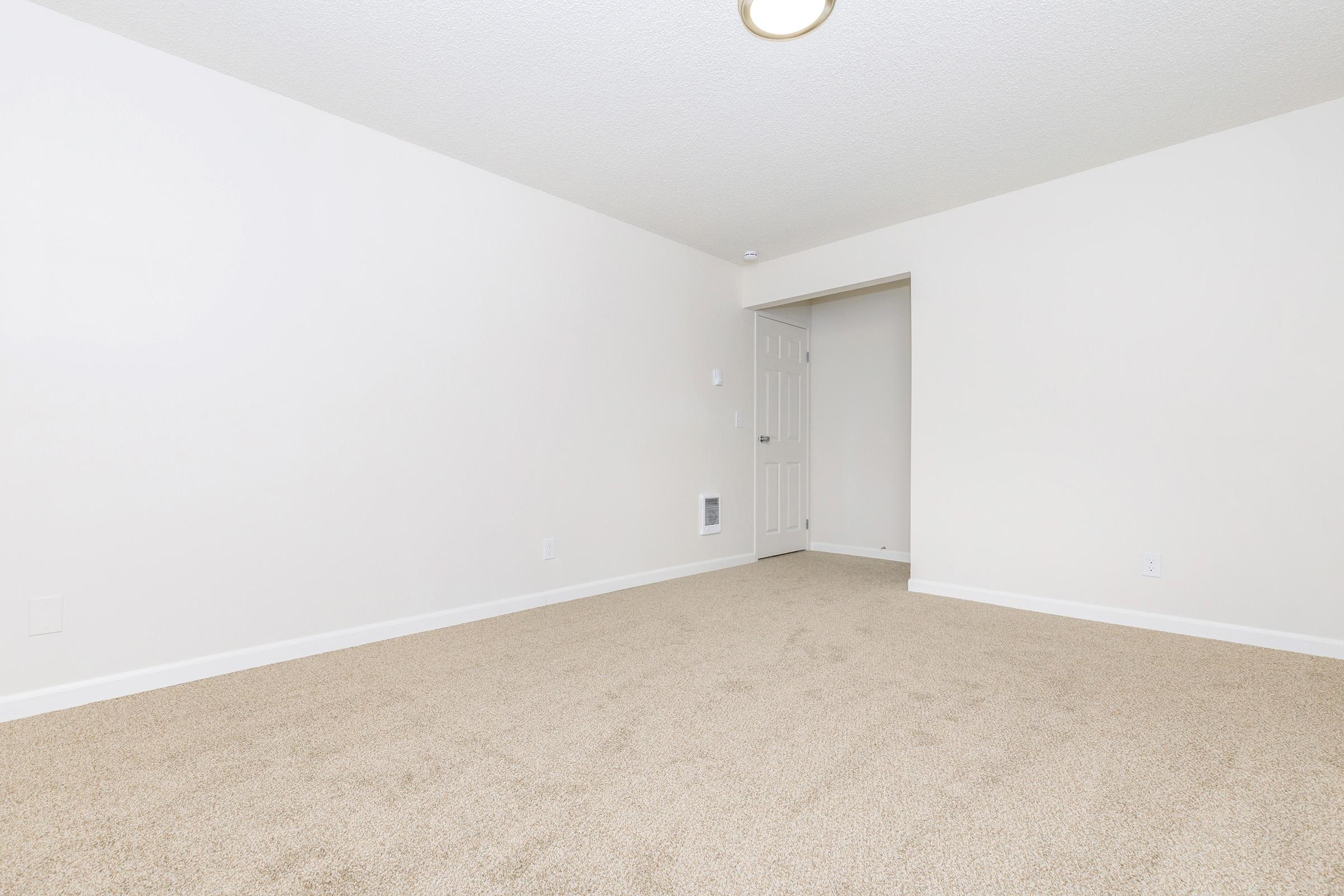 A spacious, empty room with light-colored walls and a ceiling light fixture. The floor is covered in beige carpet, and there is a doorway leading to another room in the background. The overall appearance is clean and minimalistic, creating a bright and airy atmosphere.