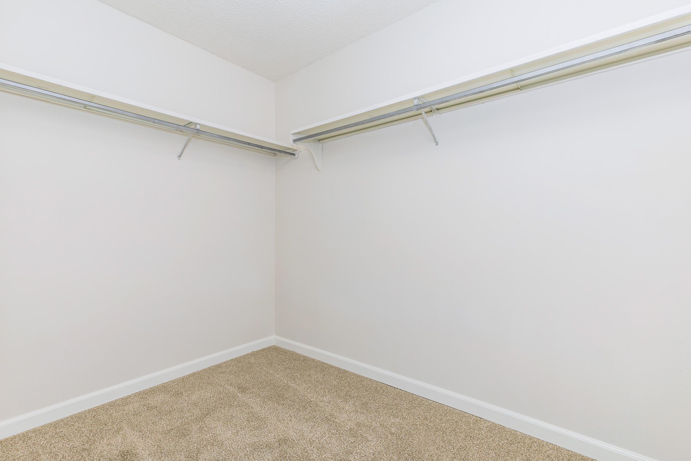 Empty closet with two metal hanging rods mounted on the walls, white walls, and a carpeted floor. The space is well-lit and uncluttered, providing a clean slate for storage or organization.
