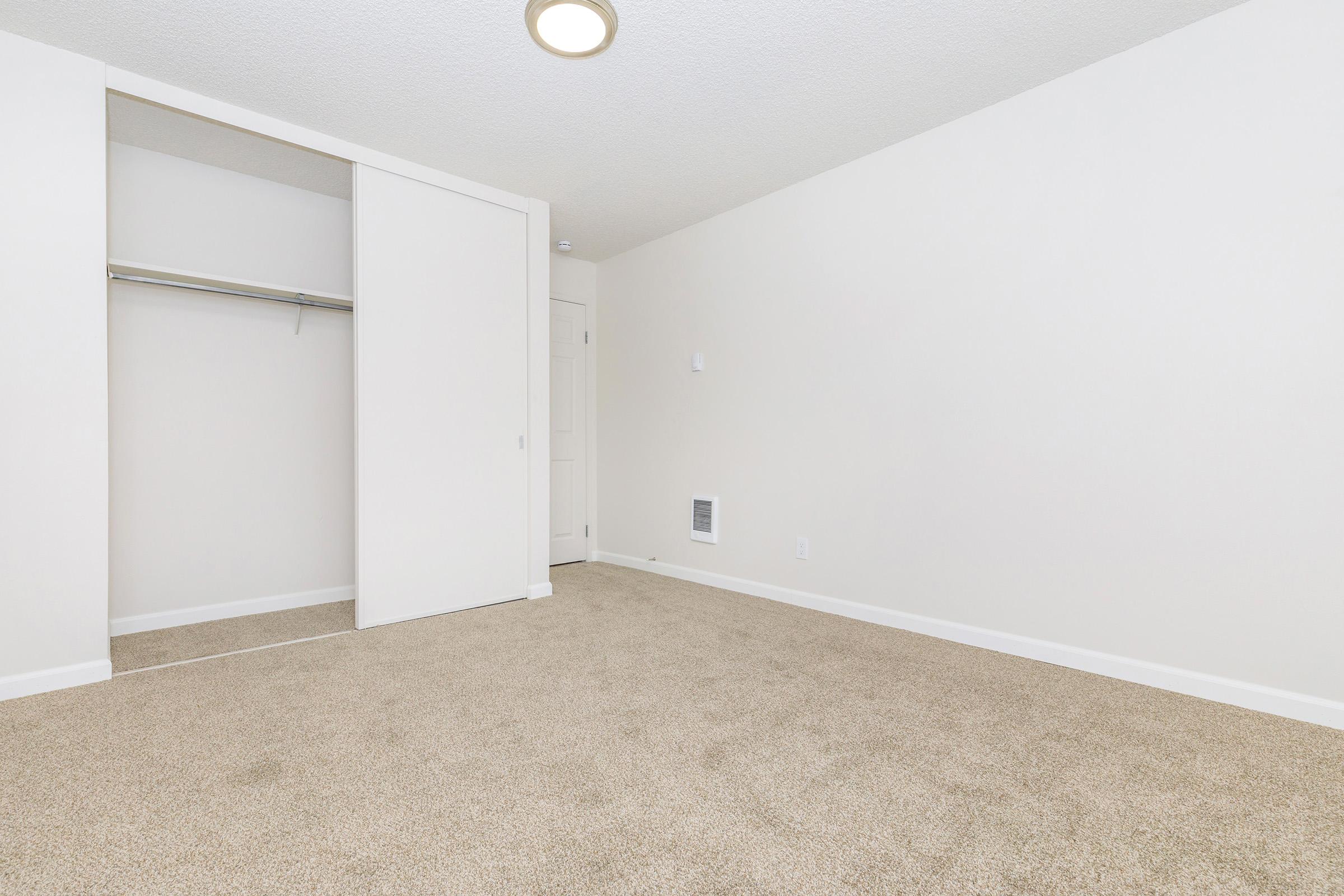 Empty room with light beige carpet, white walls, and a closet with sliding doors on one side. There is a small air vent on the wall and a ceiling light fixture. The room is spacious and bright, with a minimalistic design and no furniture.