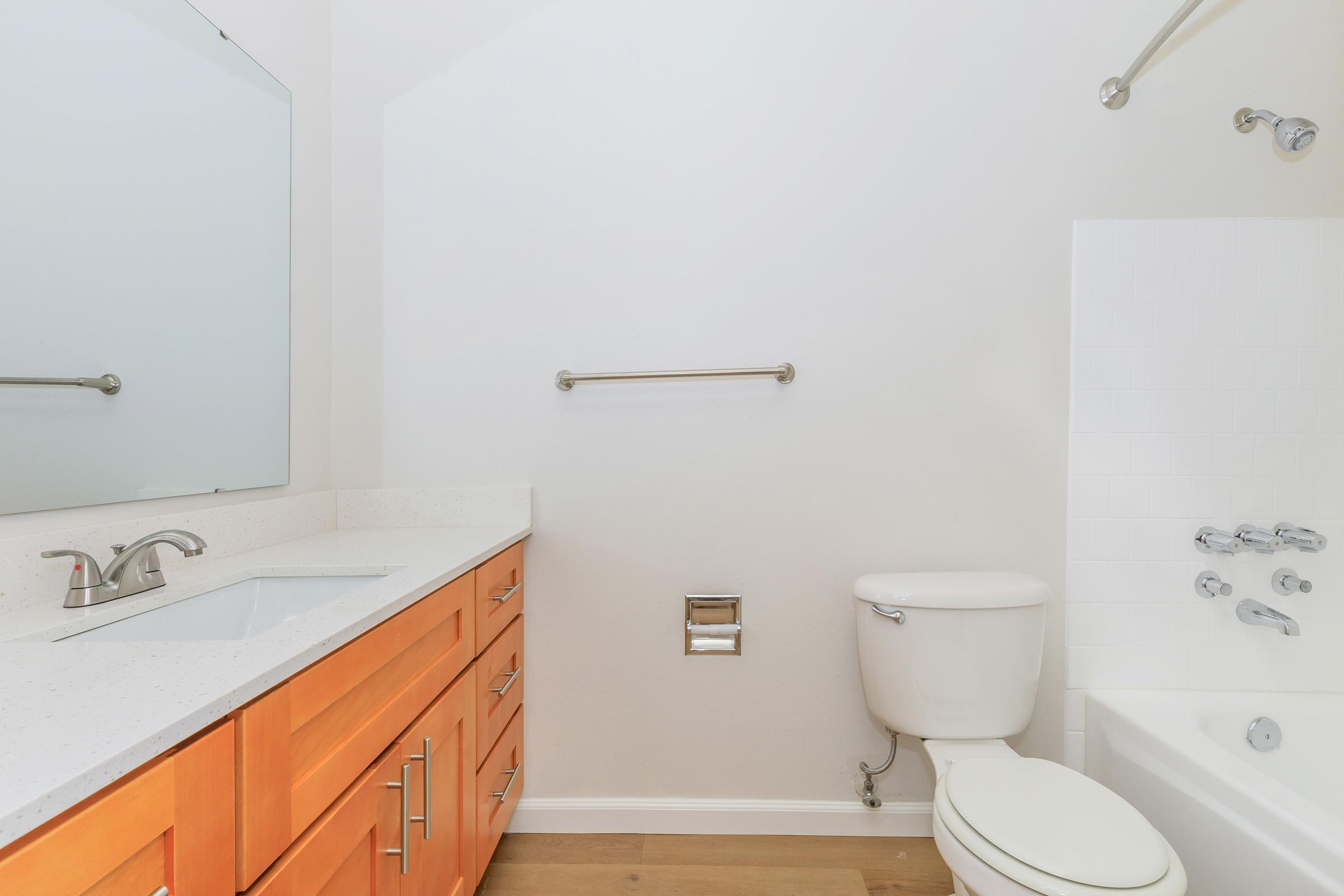 A clean and modern bathroom featuring a white tub, toilet, and an orange wood vanity. The vanity has a white countertop and a mirror above it. A towel bar is mounted on the wall, and the flooring is a light wood finish, creating a bright and spacious atmosphere.