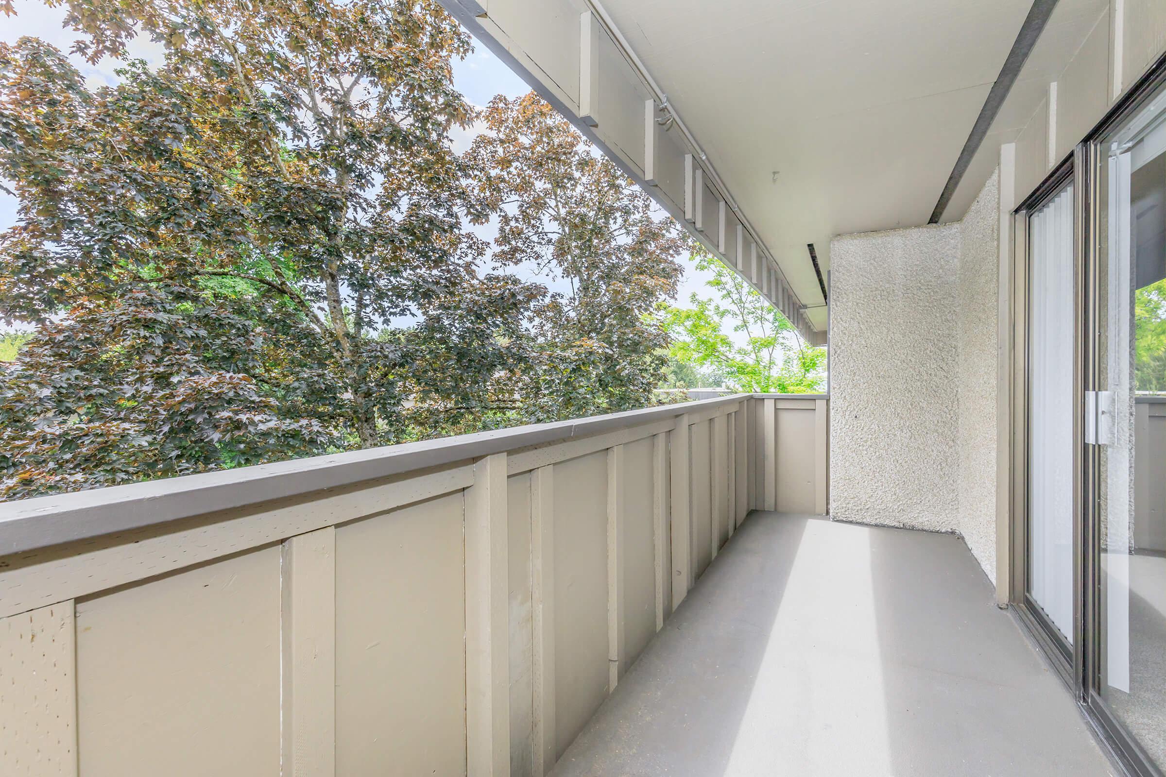 A spacious balcony with a light gray floor and beige railing, surrounded by lush green trees. The balcony is part of a modern building, featuring a smooth, clean design with ample natural light. The scene conveys a tranquil outdoor space ideal for relaxation.