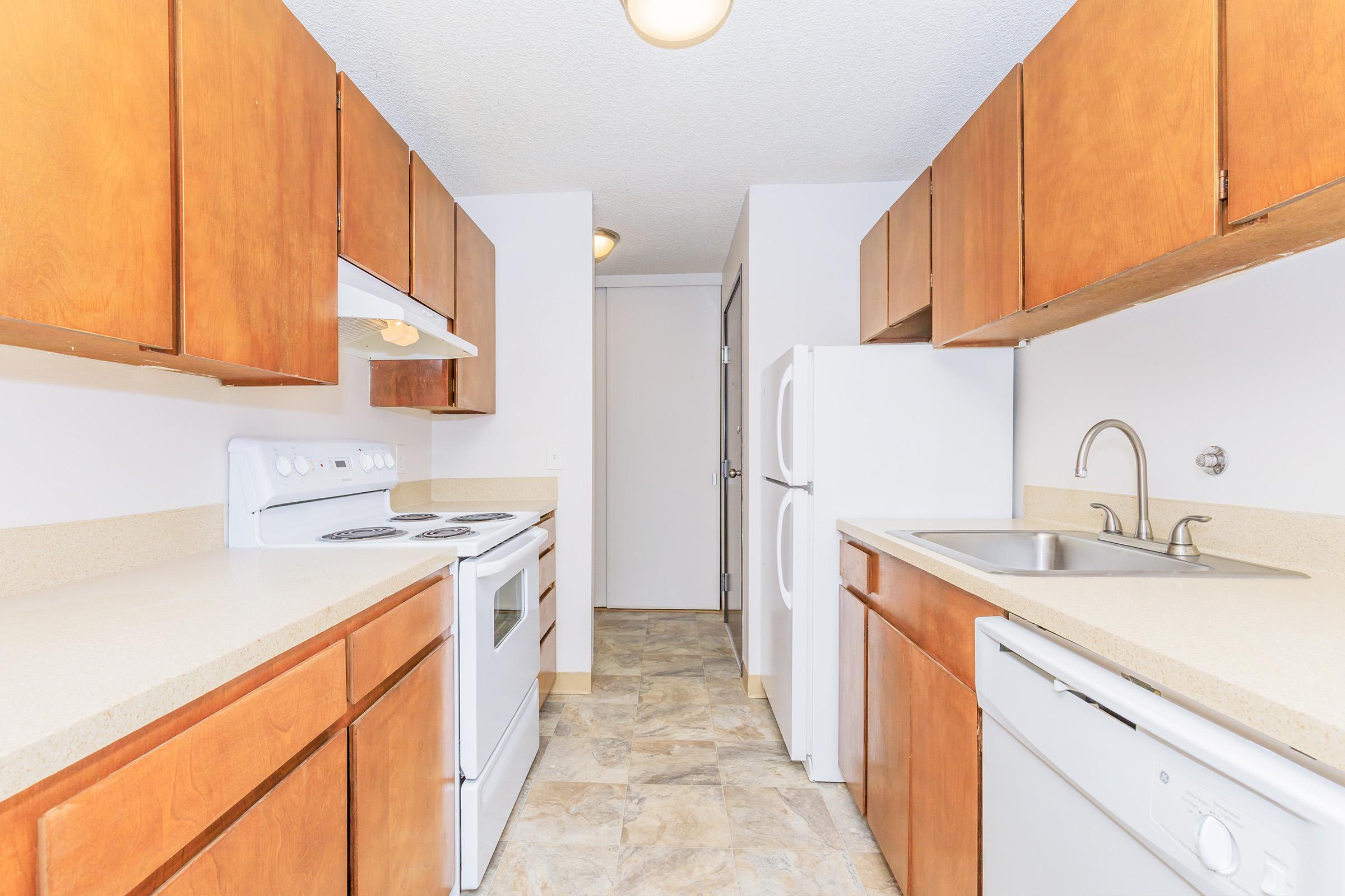 Modern kitchen interior featuring wooden cabinetry, white appliances including a stove, refrigerator, and dishwasher. Beige countertops and tiled flooring provide a clean, functional space. There is a doorway leading to another room in the background, with ample natural light illuminating the area.