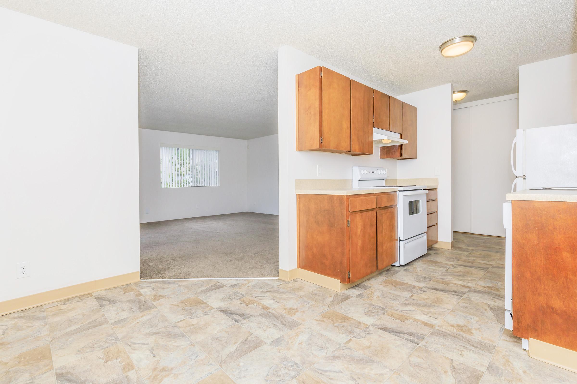 Spacious apartment interior featuring a kitchen with brown wooden cabinets and white appliances. A living area is visible in the background with natural light coming through a window. The floor has a tiled design, and the walls are painted white, creating a bright and open atmosphere.