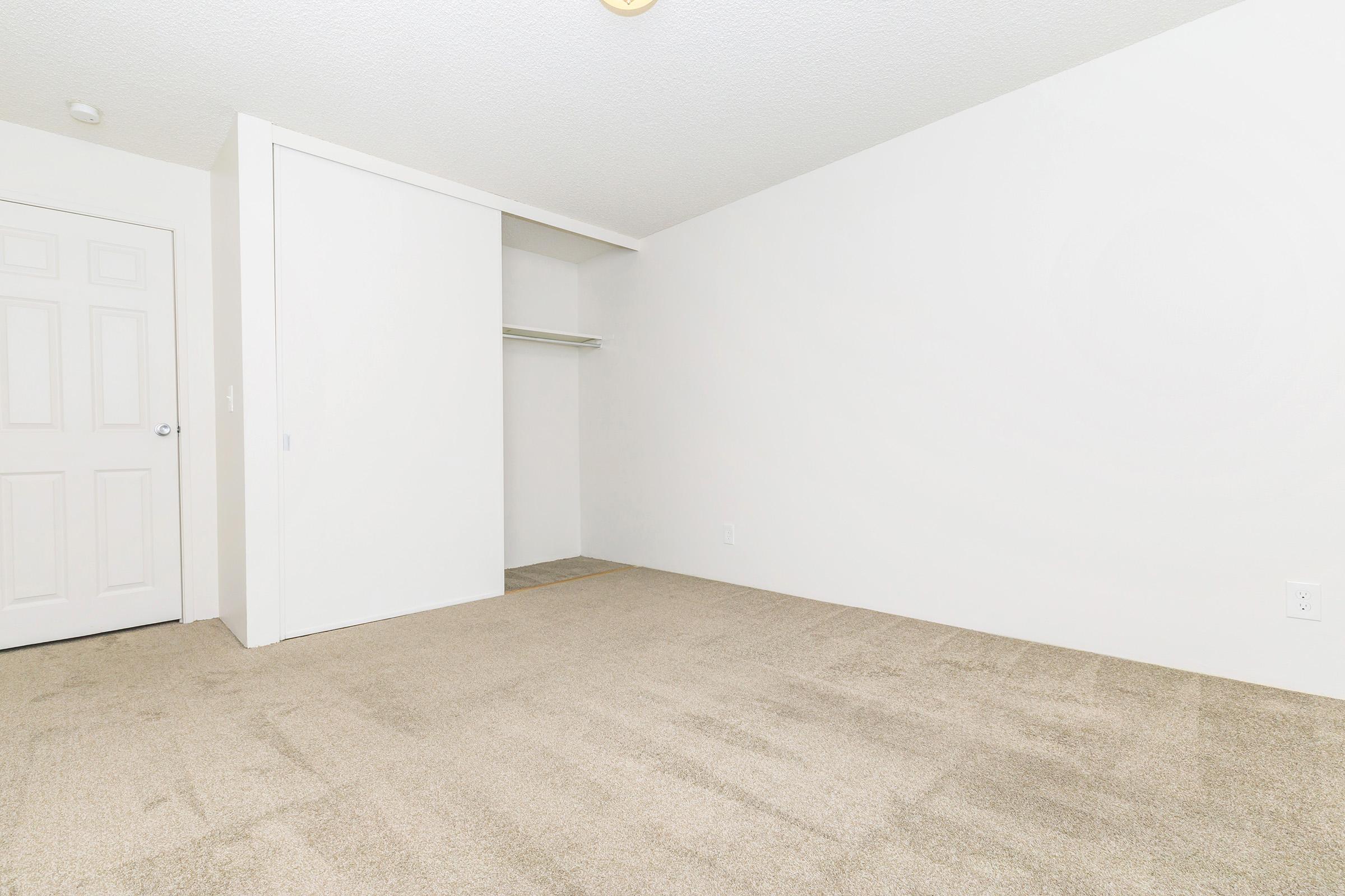 A spacious, empty room with light-colored walls and a beige carpet. There is a closet with sliding doors on the left side, and the room features a ceiling light fixture. The overall atmosphere is bright and minimalistic, ideal for various uses.