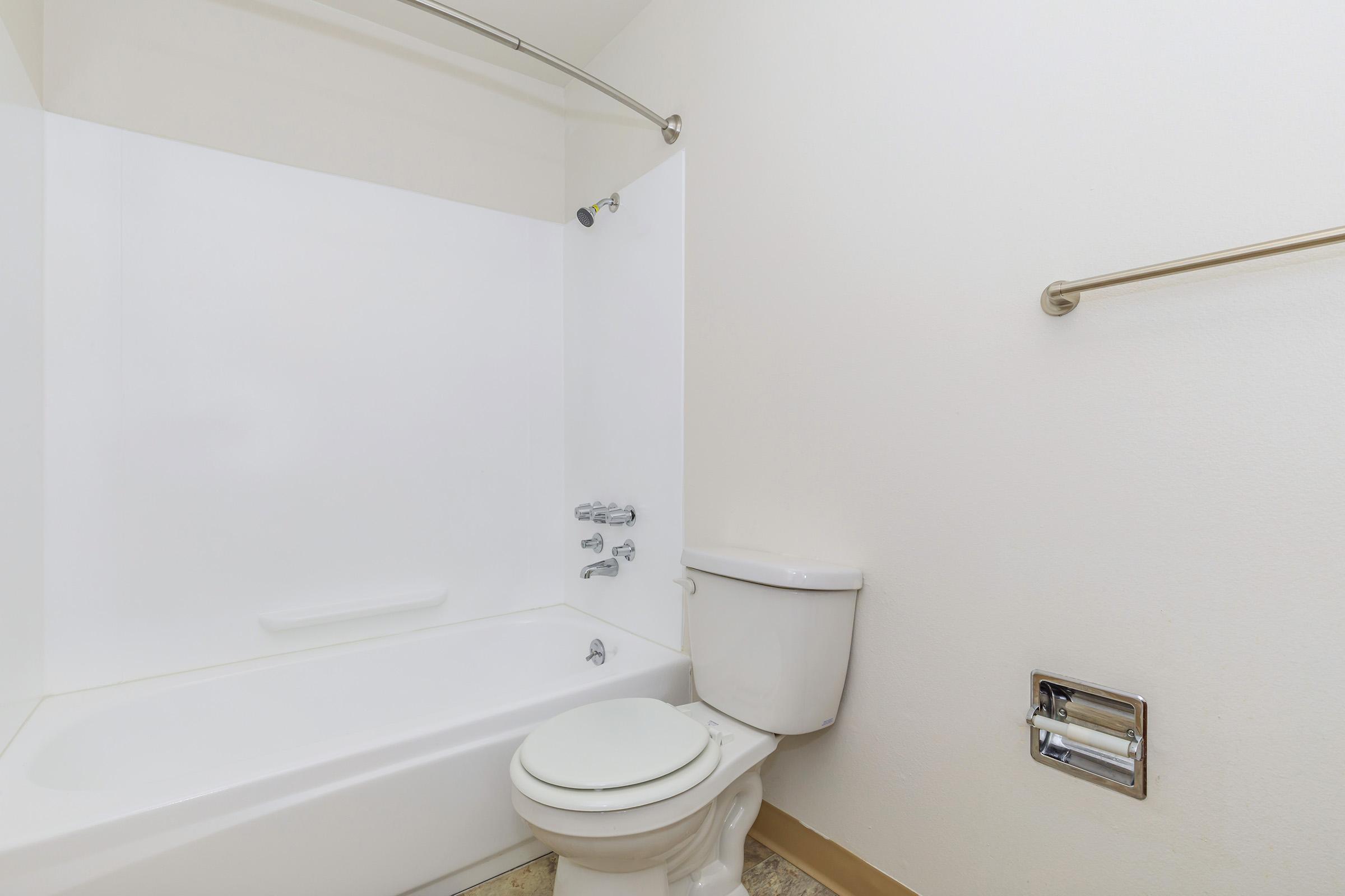 A clean and minimalistic bathroom featuring a white bathtub with a showerhead, a white toilet, and a towel rack. The walls are light-colored, and the floor has a simple tile design. The overall appearance is bright and spacious, creating a fresh and modern atmosphere.