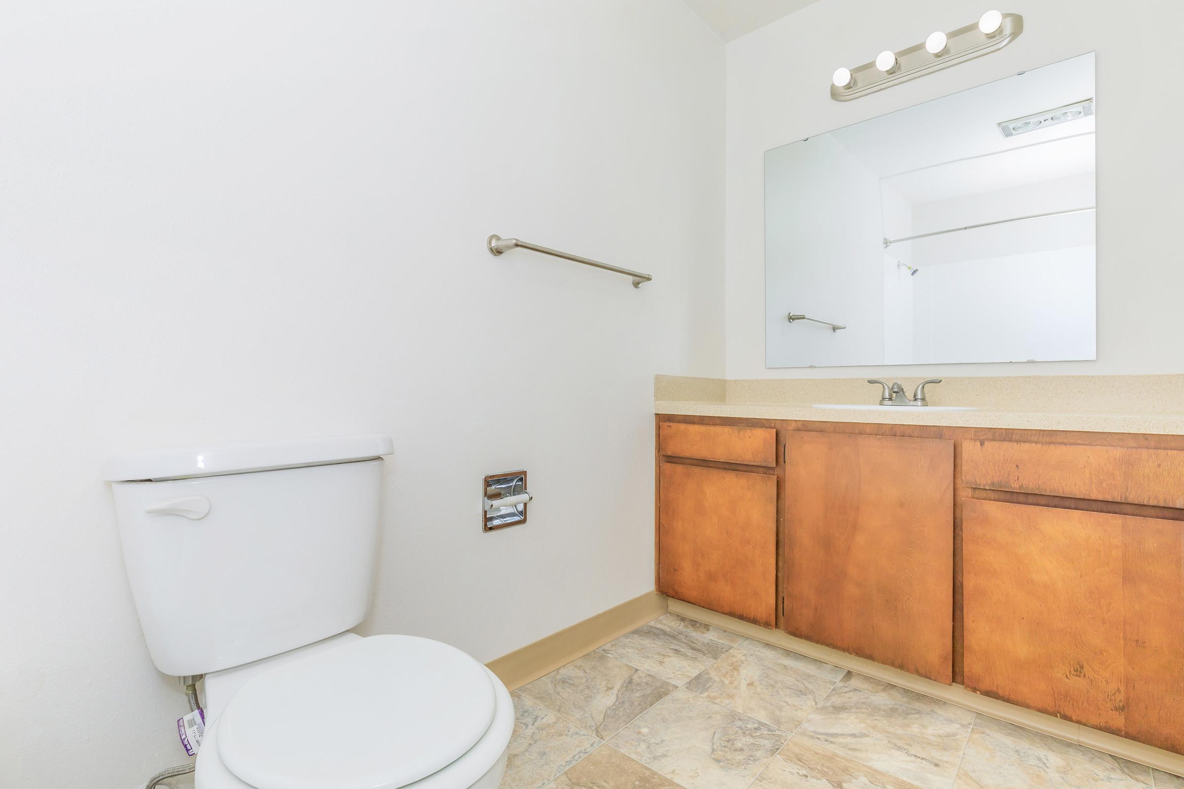 A clean and modern bathroom featuring a white toilet, wooden cabinets under a sink, a large mirror, and a light fixture above. The floor is tiled with a neutral stone pattern, and the walls are painted white, creating a bright and simple aesthetic.