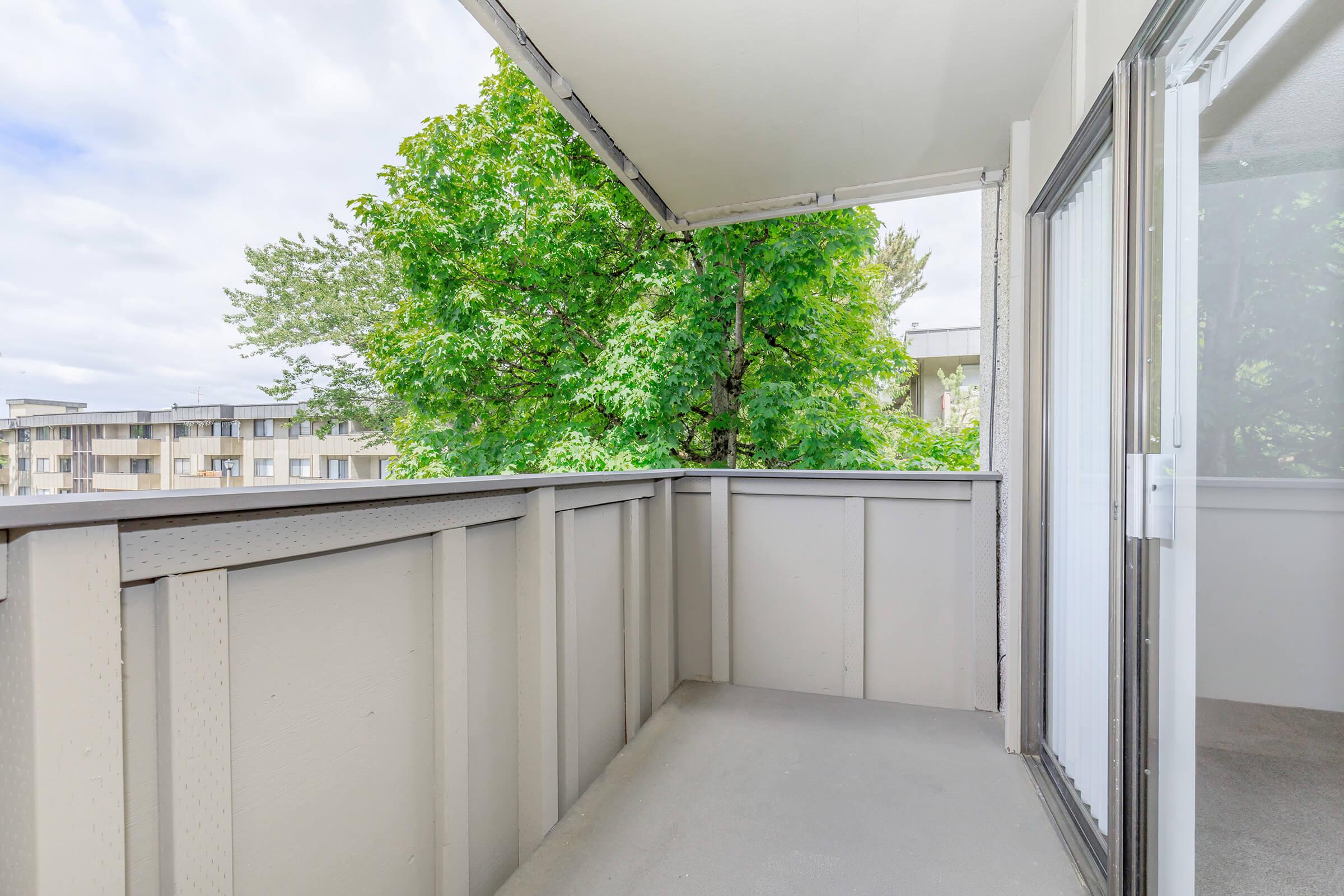 A spacious balcony or porch with a light gray floor, surrounded by greenery from trees. The view includes a residential building in the background and a cloudy sky, creating a serene outdoor space for relaxation or recreation.