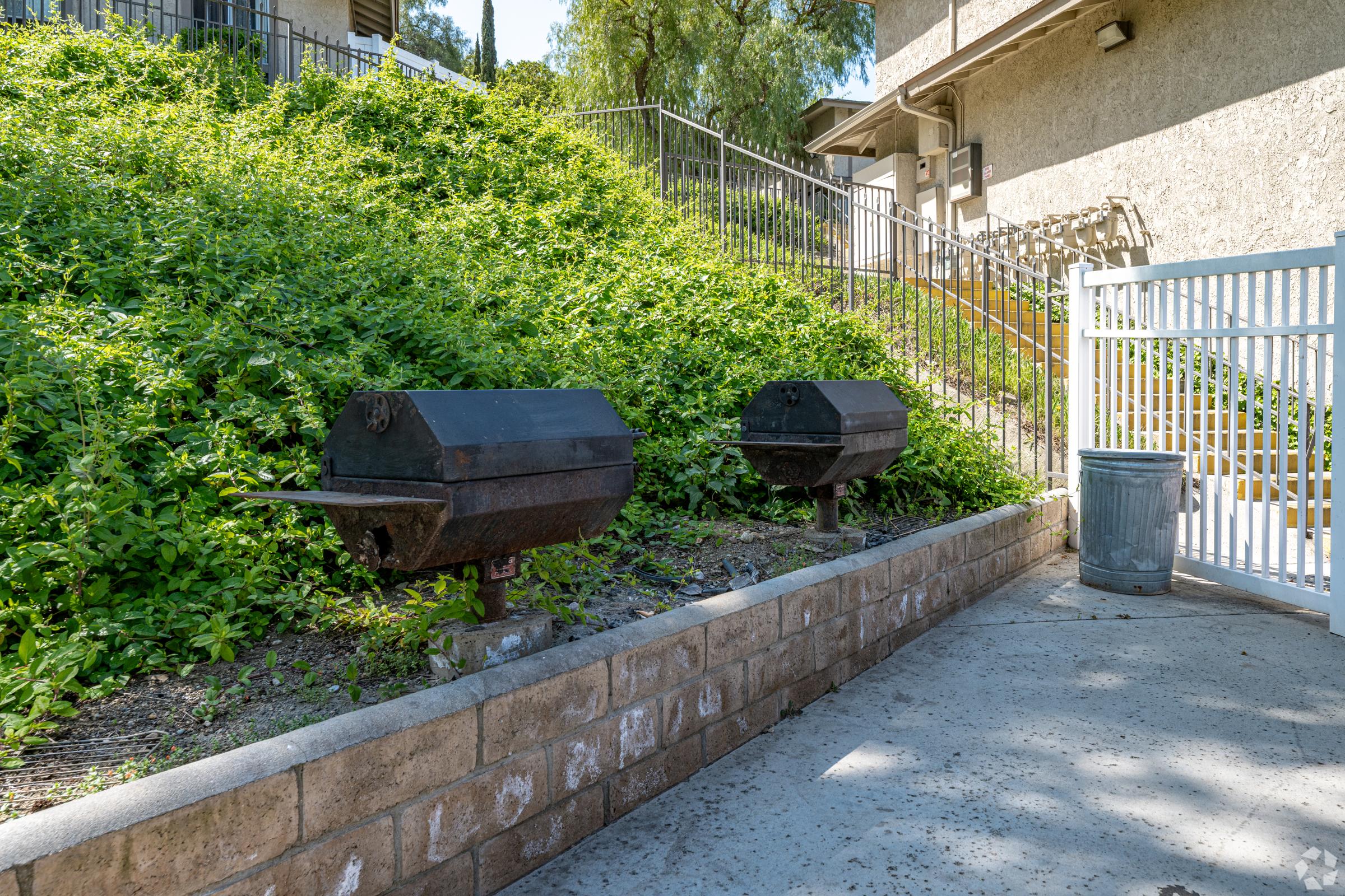 Two black charcoal grills positioned on a stone ledge beside a grassy hill, with a trash can nearby. The area appears well-lit and is surrounded by green foliage and a white fence, indicating a communal outdoor space.