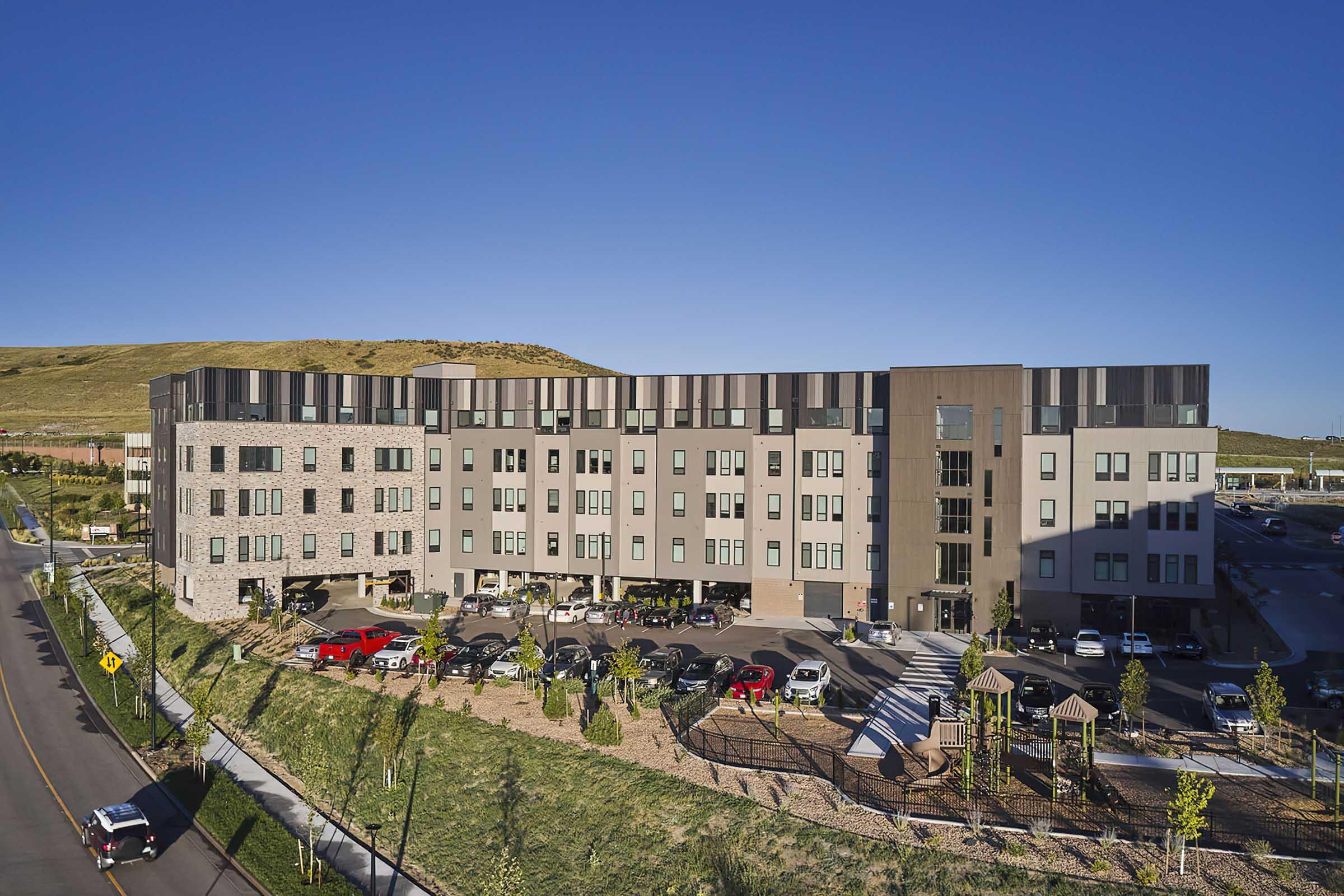 A modern multi-story building with a mix of stone and metal exterior, surrounded by greenery and parked cars. The setting features a clear blue sky and a hillside in the background, indicating a suburban area. The architecture showcases contemporary design elements.