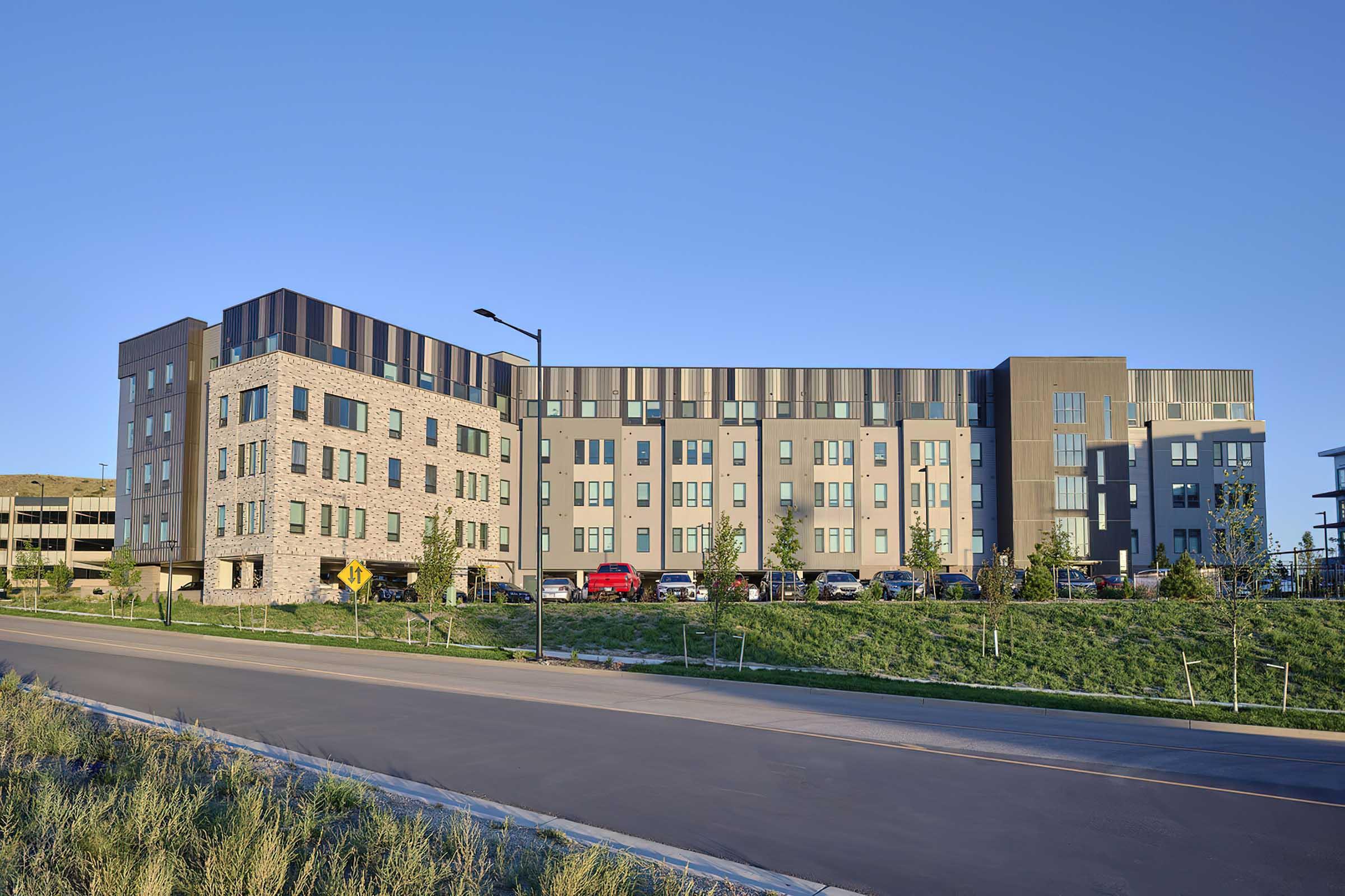 Modern multi-story building featuring a mix of stone and sleek metal exteriors. The structure is flanked by landscaped green spaces and a paved road. Multiple vehicles are parked in front, suggesting an active commercial or residential space in a suburban setting. Clear blue sky serves as the backdrop.