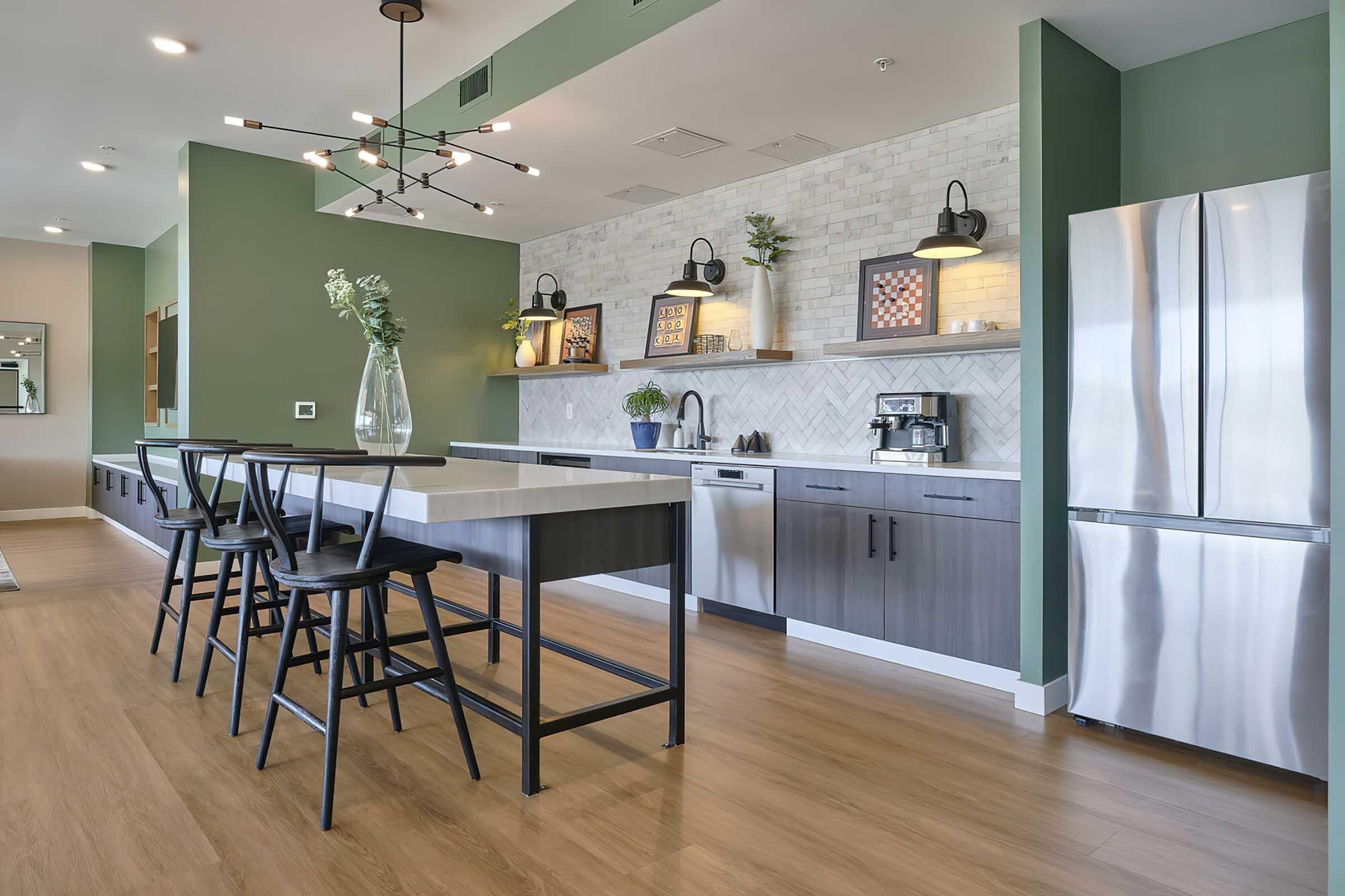 Modern kitchen featuring a large island with seating, sleek cabinetry, and stainless steel appliances. The walls are painted in a soft green, and decorative lighting fixtures hang above the island. Decorative elements like plants and artwork add a stylish touch to the space. Wood flooring completes the contemporary look.