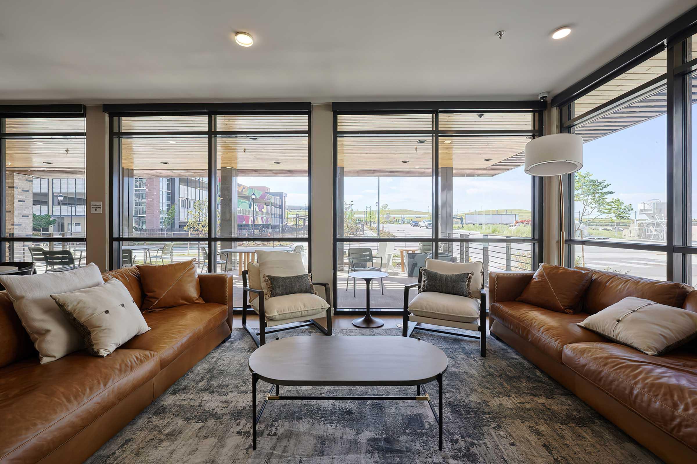 A modern living area featuring two brown leather sofas with decorative pillows, a coffee table in the center, and large windows allowing natural light. The view outside includes tables and chairs on a patio, with greenery visible in the distance. The decor is contemporary and inviting.