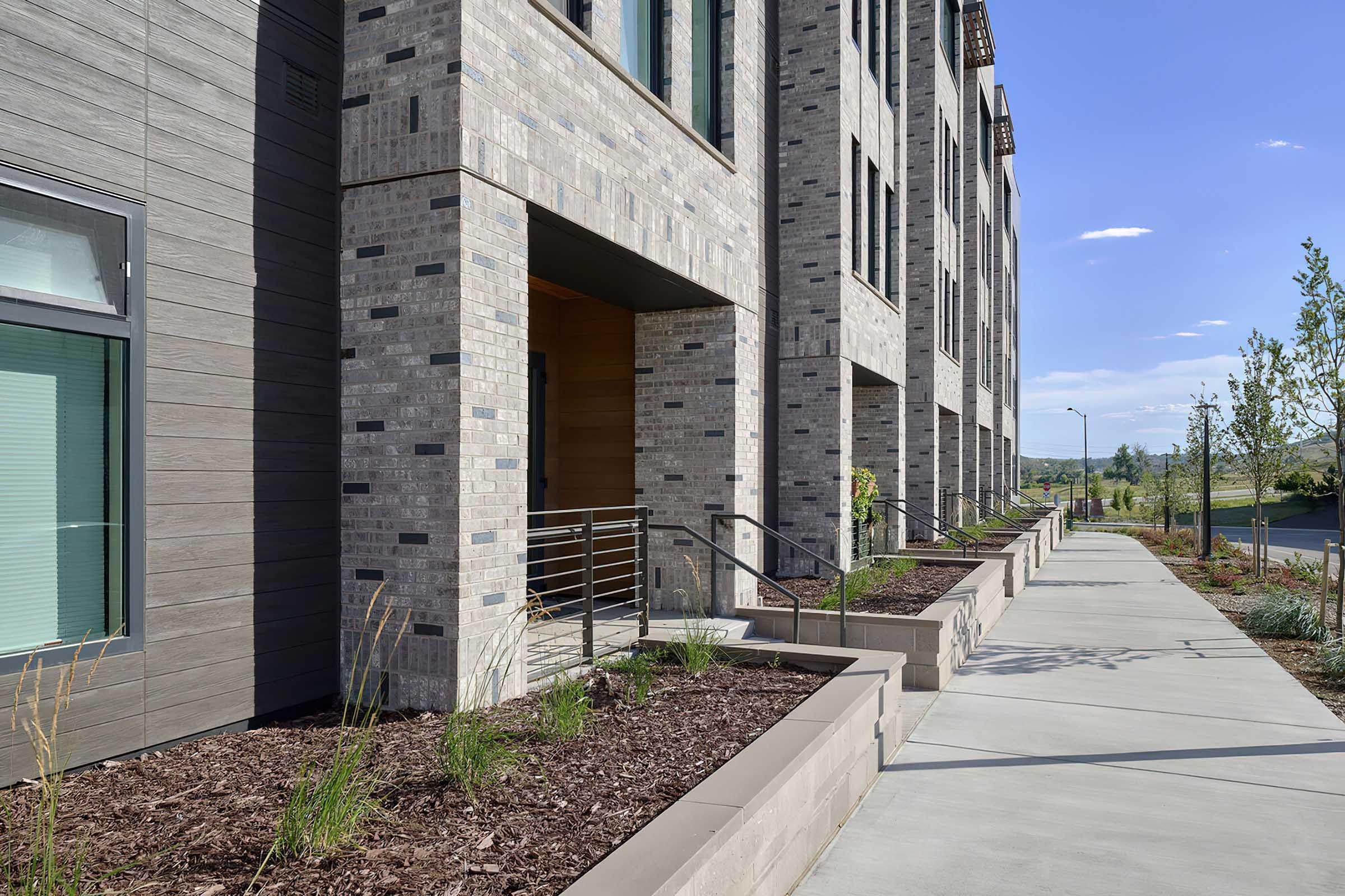 Modern architectural design featuring a row of multi-story buildings with brick and concrete façades. The sidewalk runs alongside landscaped planters filled with plants, and there are entrances with metal railings. The scene is set under a clear blue sky.