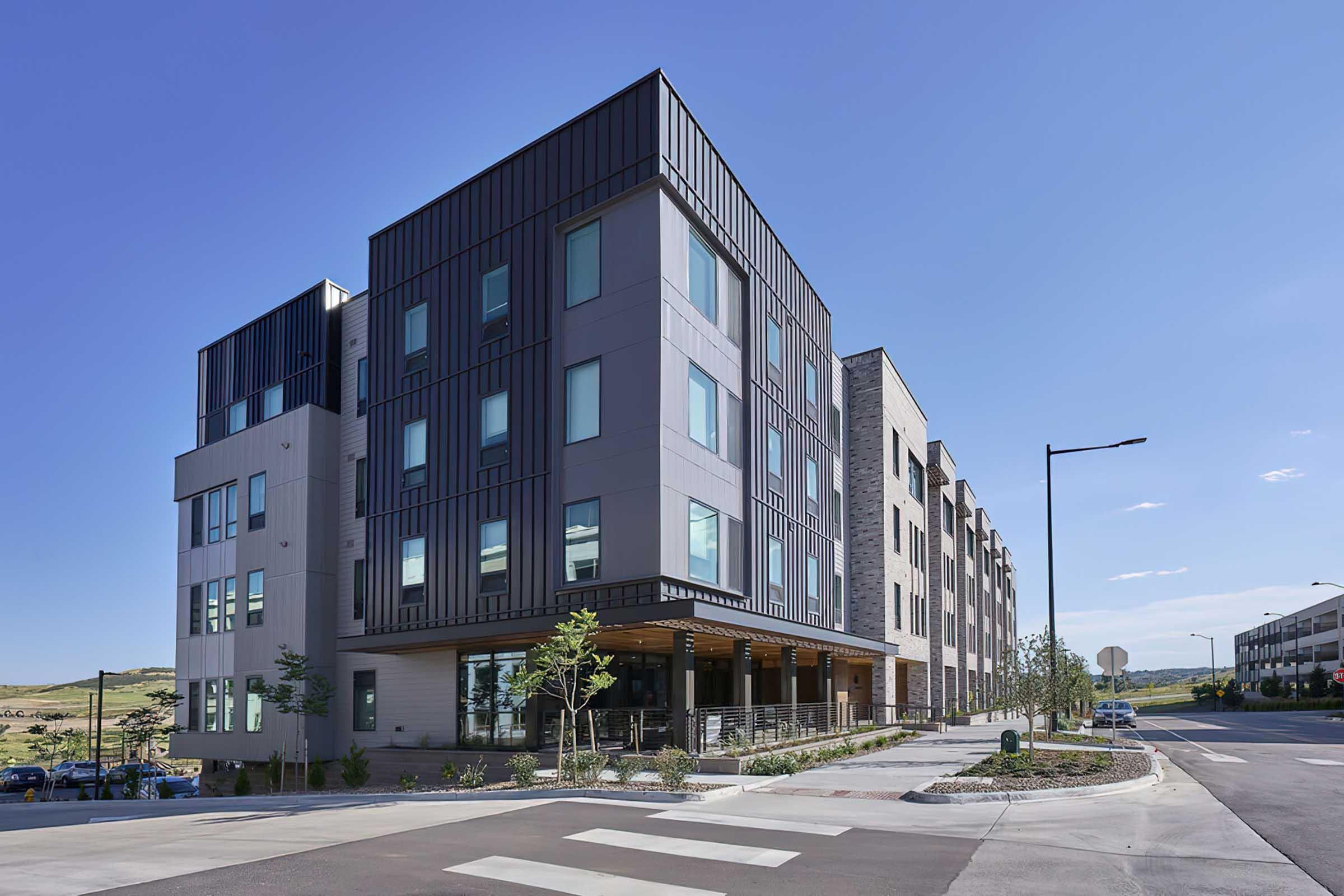 Modern multi-story building featuring a sleek design with large windows, integrated greenery, and a mix of materials. Located on a wide street with clear skies and surrounding landscape, the structure represents contemporary architecture in an urban setting. The entrance is highlighted with steps and landscaping.