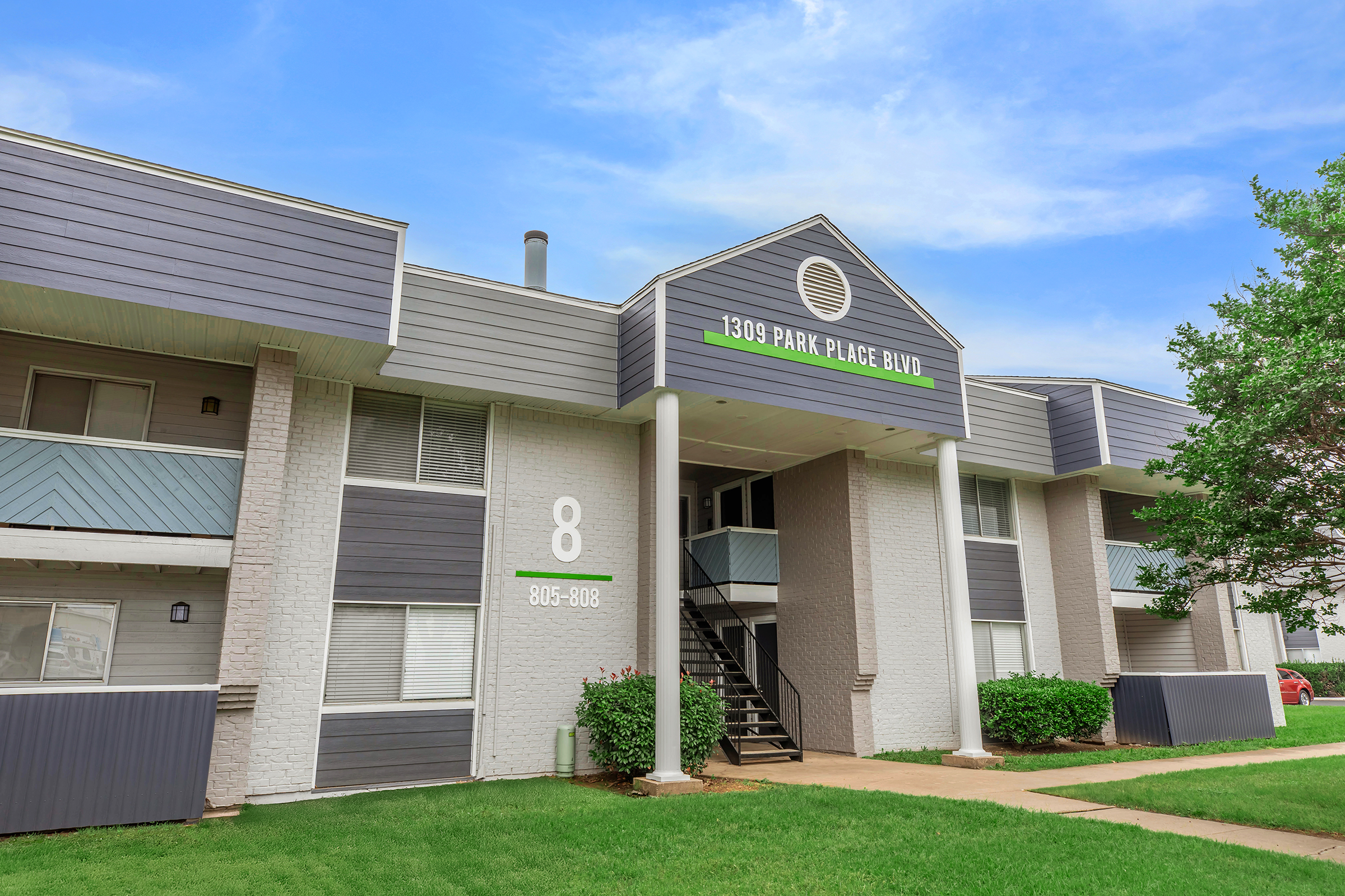 Exterior view of a residential building with a light gray facade and a peaked roof. The entrance features a staircase leading to the upper level, and the address "1309 PARK PLACE BLVD" is displayed prominently. Green grass surrounds the building, and the sky is bright with scattered clouds.