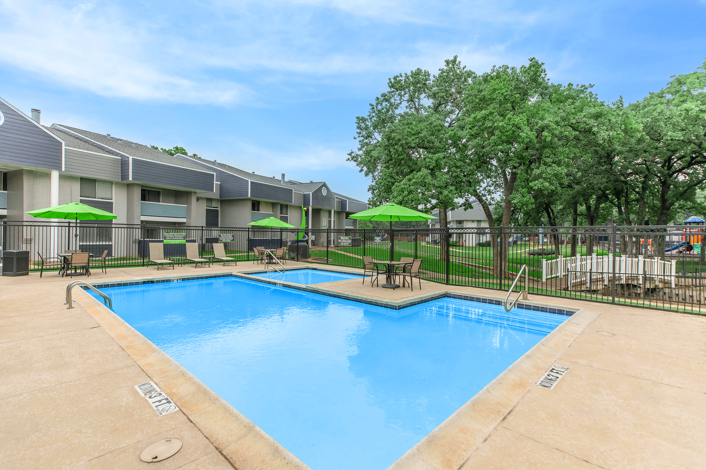 A clear, blue swimming pool surrounded by a tan concrete deck. Green umbrellas are set up over tables and chairs near the pool. In the background, there are gray apartment buildings and large green trees under a clear blue sky, creating a relaxing outdoor environment.