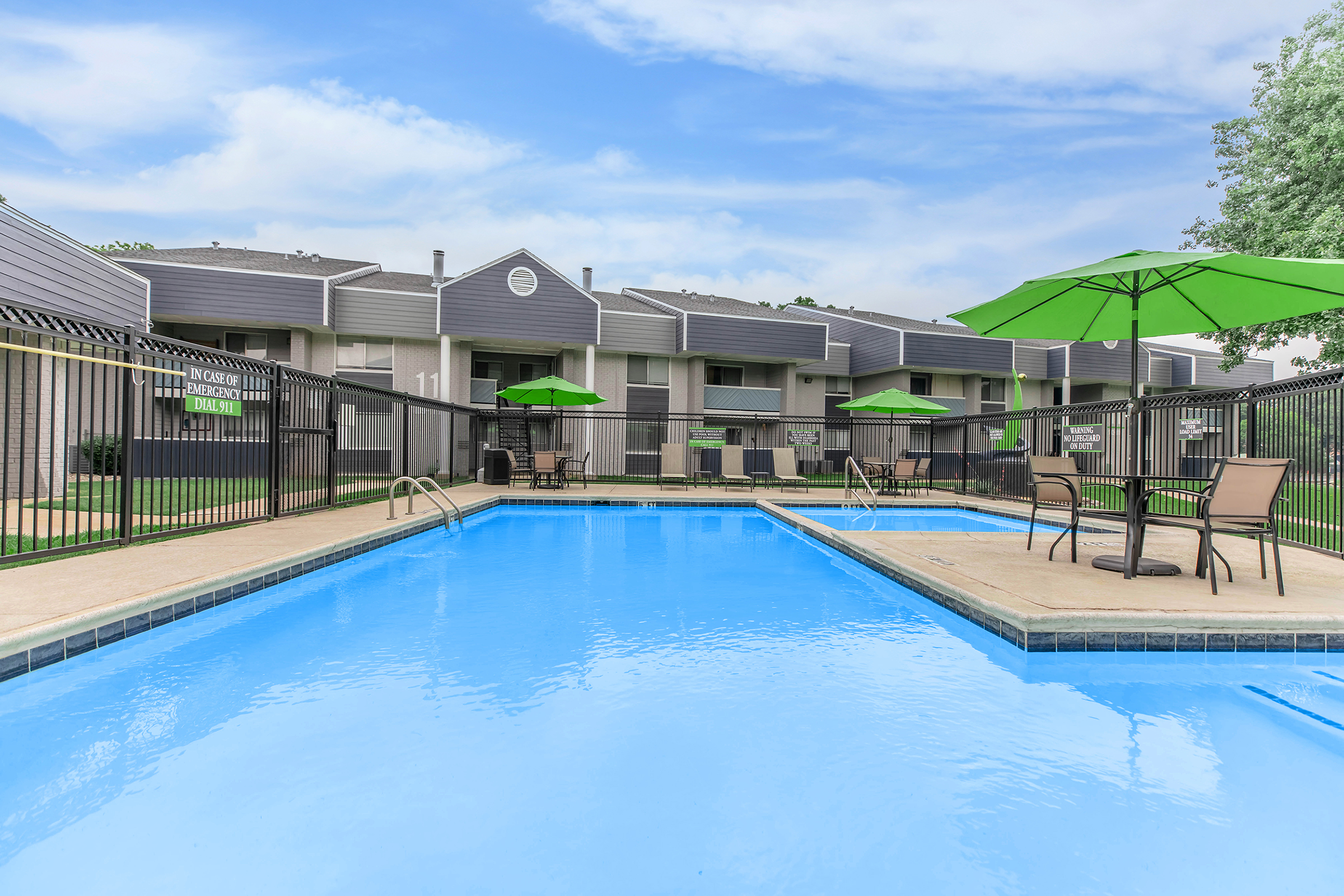 A clean swimming pool surrounded by green umbrellas and lounge chairs. In the background, there are modern apartment buildings with gray exteriors. The sky is clear with a few clouds, creating a pleasant and inviting atmosphere.