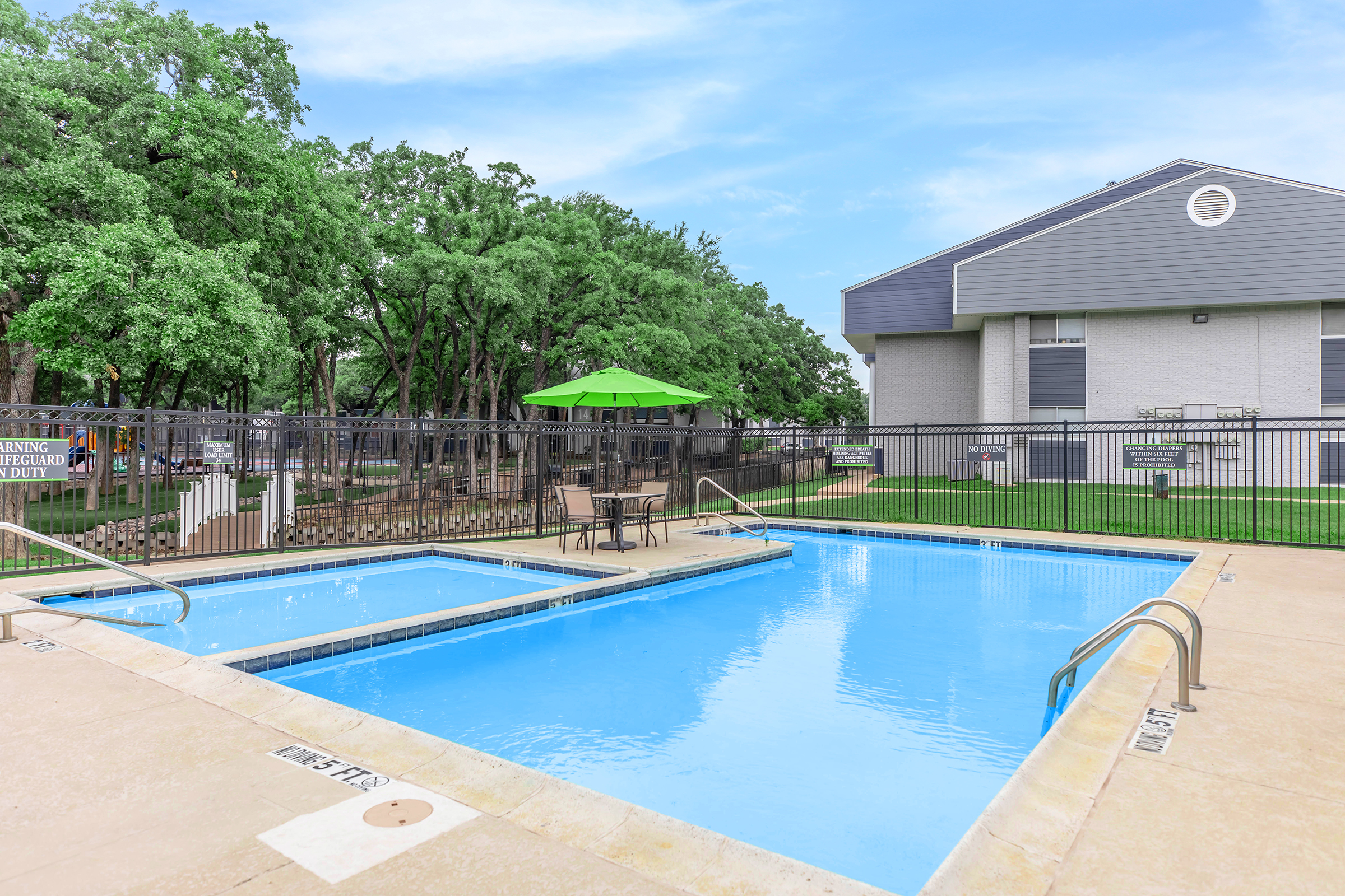 A clean, inviting swimming pool with a smaller hot tub adjacent to it, surrounded by a paved area. In the background, there are trees providing shade and a green umbrella on a table. A building is visible to the right, indicating a residential area. The sky is clear with a hint of clouds.