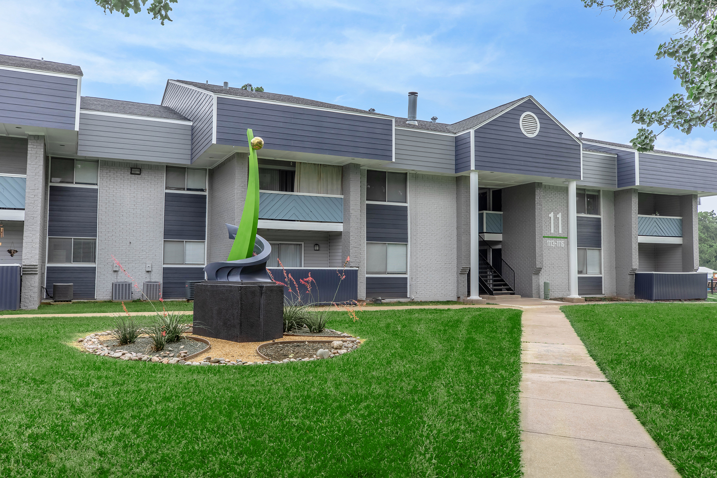 A contemporary apartment building featuring a unique green sculpture in a landscaped area. The building has a mix of gray and light-colored exteriors, with a walkway leading to the entrance marked "11." Surrounding greenery includes grass and ornamental plants, creating a welcoming environment.