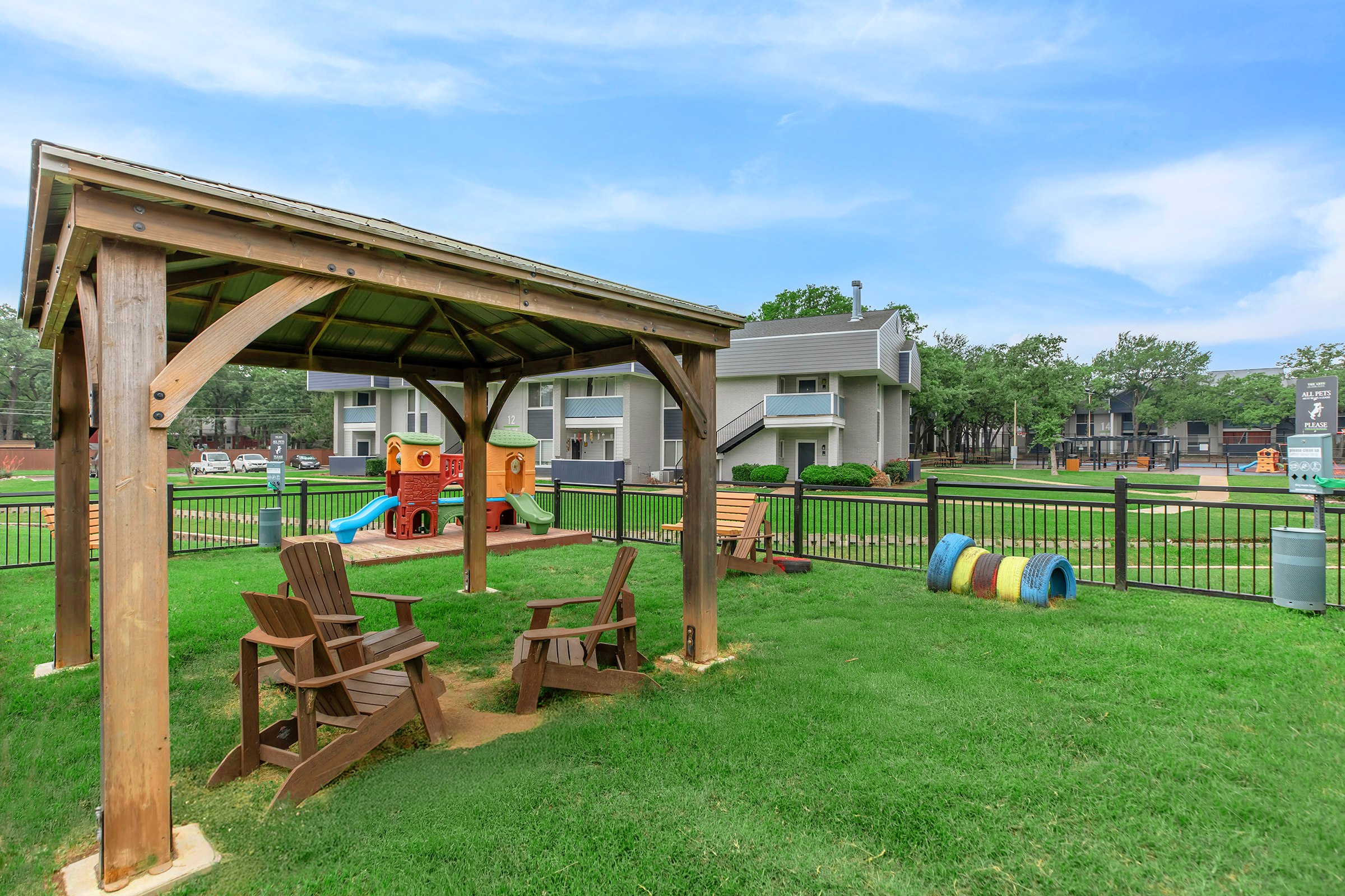 A wooden gazebo with seating overlooks a playground with a colorful slide and play equipment. Surrounding the area is green grass and a fenced yard, with residential buildings visible in the background under a blue sky.