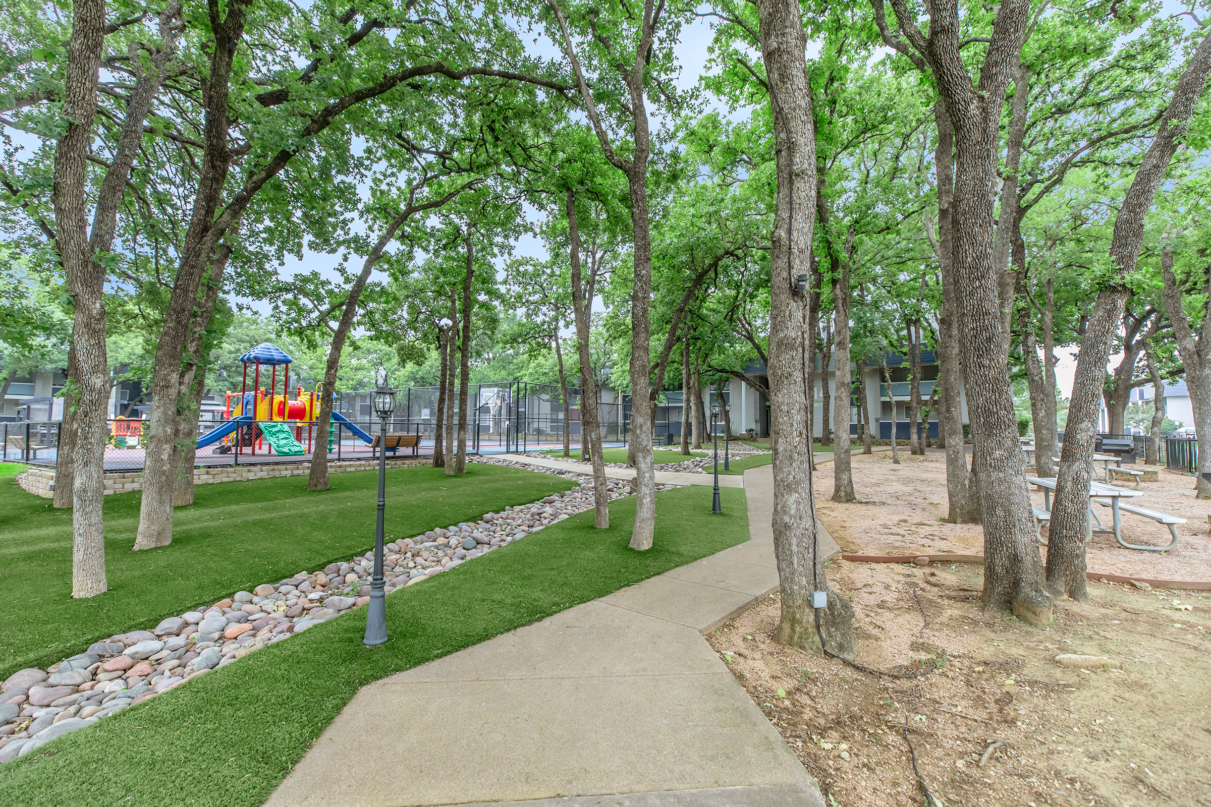 A landscaped park area featuring green grass, a playground with colorful equipment, and a walking path lined with trees. Picnic tables are visible near a sandy area, with decorative stones along the path. The scene is bright and inviting, suitable for outdoor activities.