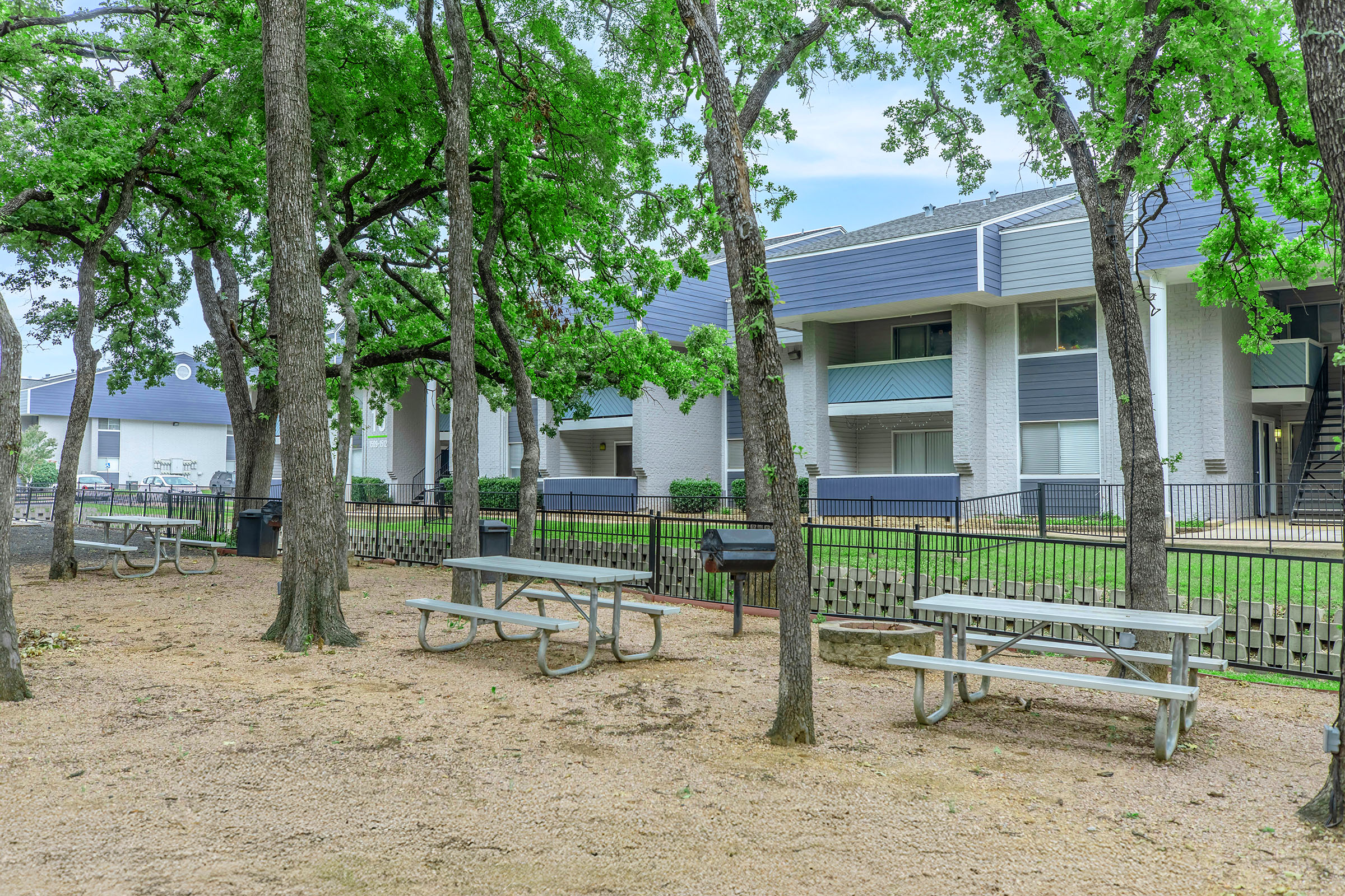 A serene outdoor area featuring several picnic tables surrounded by trees. A charcoal grill is visible nearby, with a view of multi-story apartment buildings in the background. The ground is sandy, and the setting appears inviting for gatherings or relaxation.