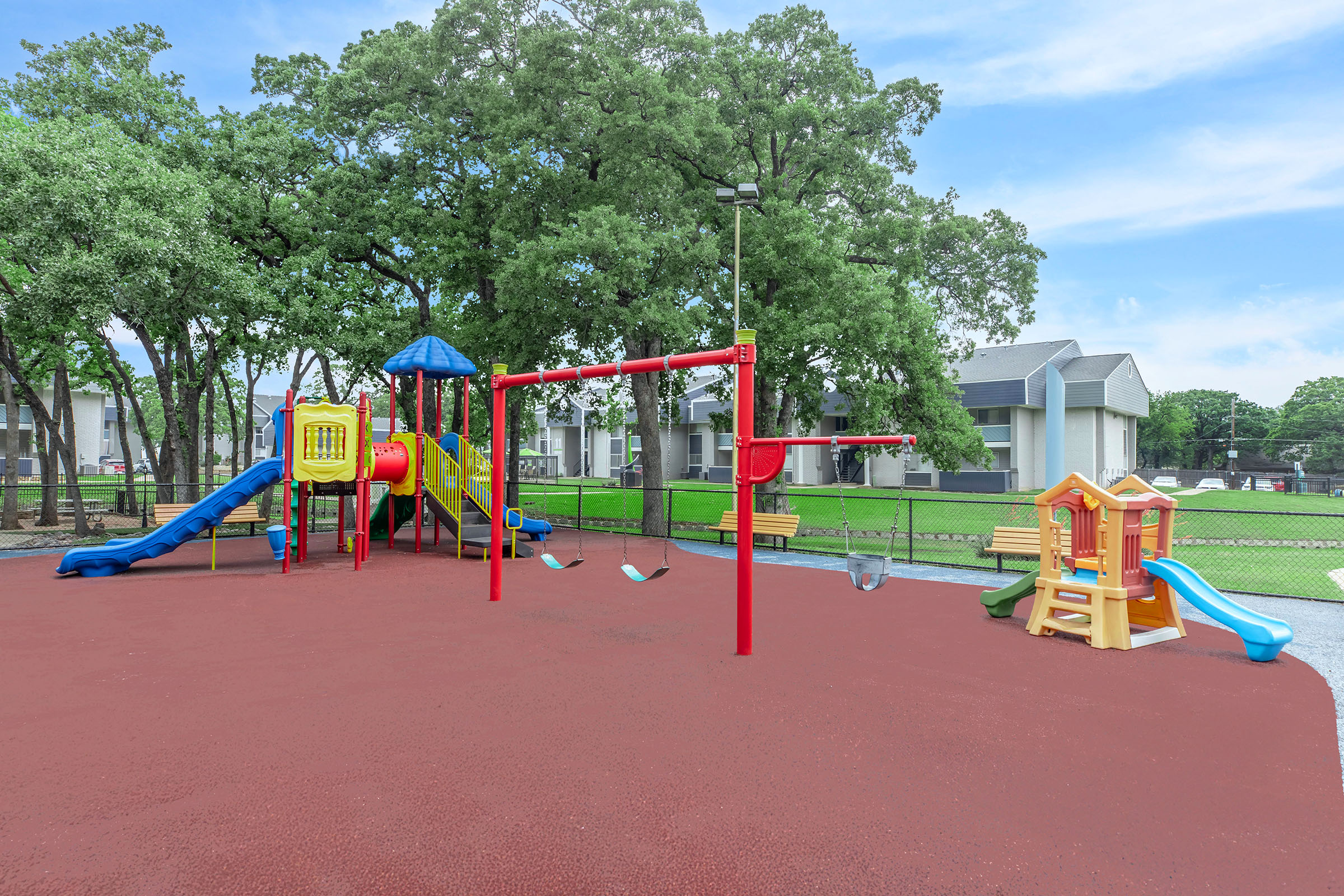 A colorful playground featuring slides, swings, climbing structures, and playhouses, set on a rubberized surface. Surrounding trees provide shade, and nearby residential buildings are visible in the background under a clear blue sky.