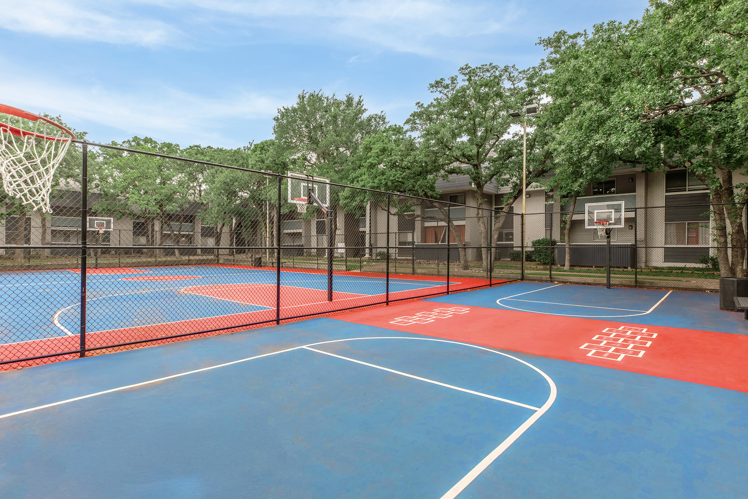 A colorful outdoor basketball court with red and blue painted surfaces. The court is enclosed by a black chain-link fence, featuring multiple basketball hoops. Surrounding trees provide shade, and there are nearby residential buildings visible in the background. The sky is clear and blue.