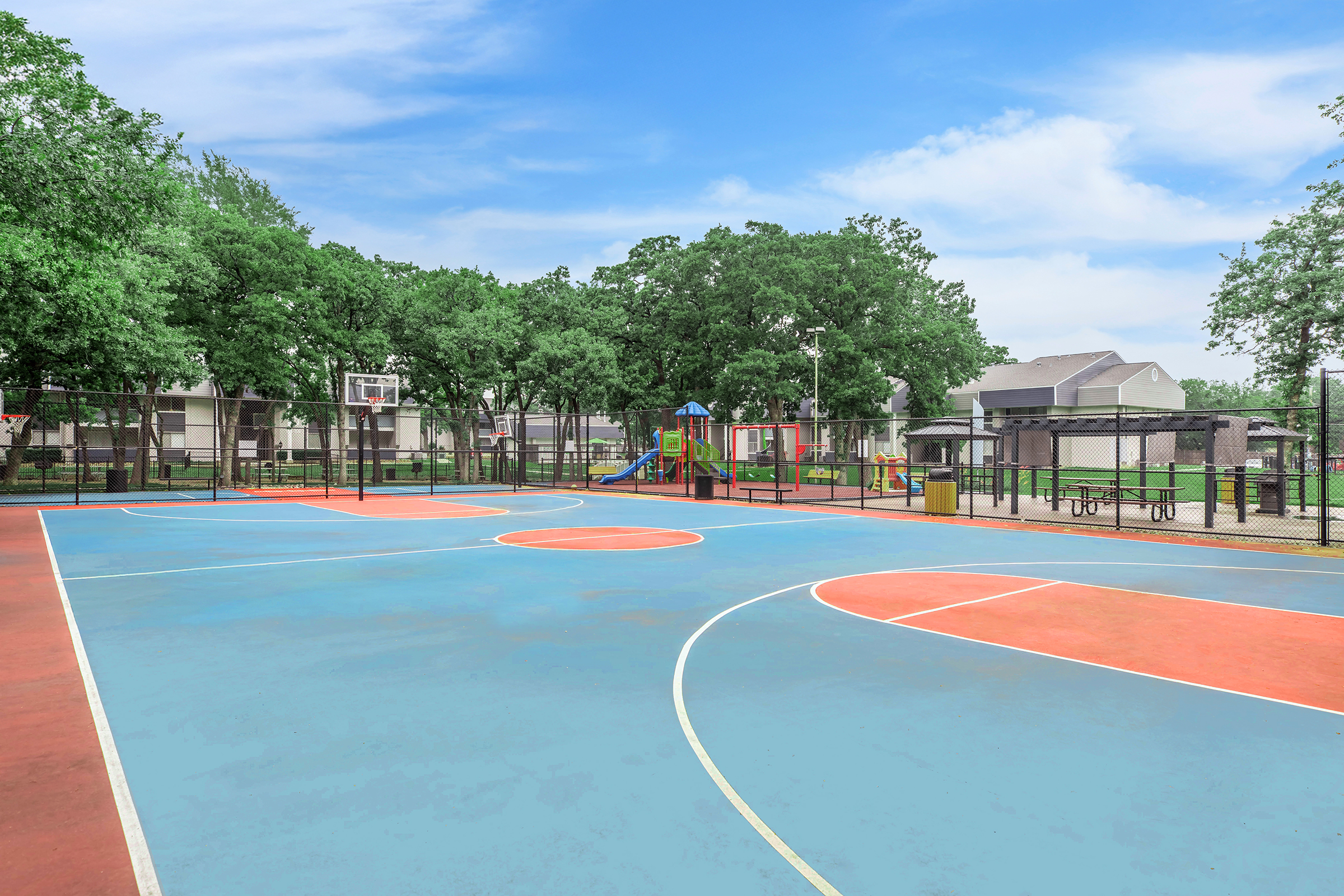 A vibrant outdoor basketball court featuring colorful blue and orange markings, surrounded by green trees. In the background, a playground with slides and climbing structures can be seen, along with several residential buildings. The sky is clear with a few clouds.