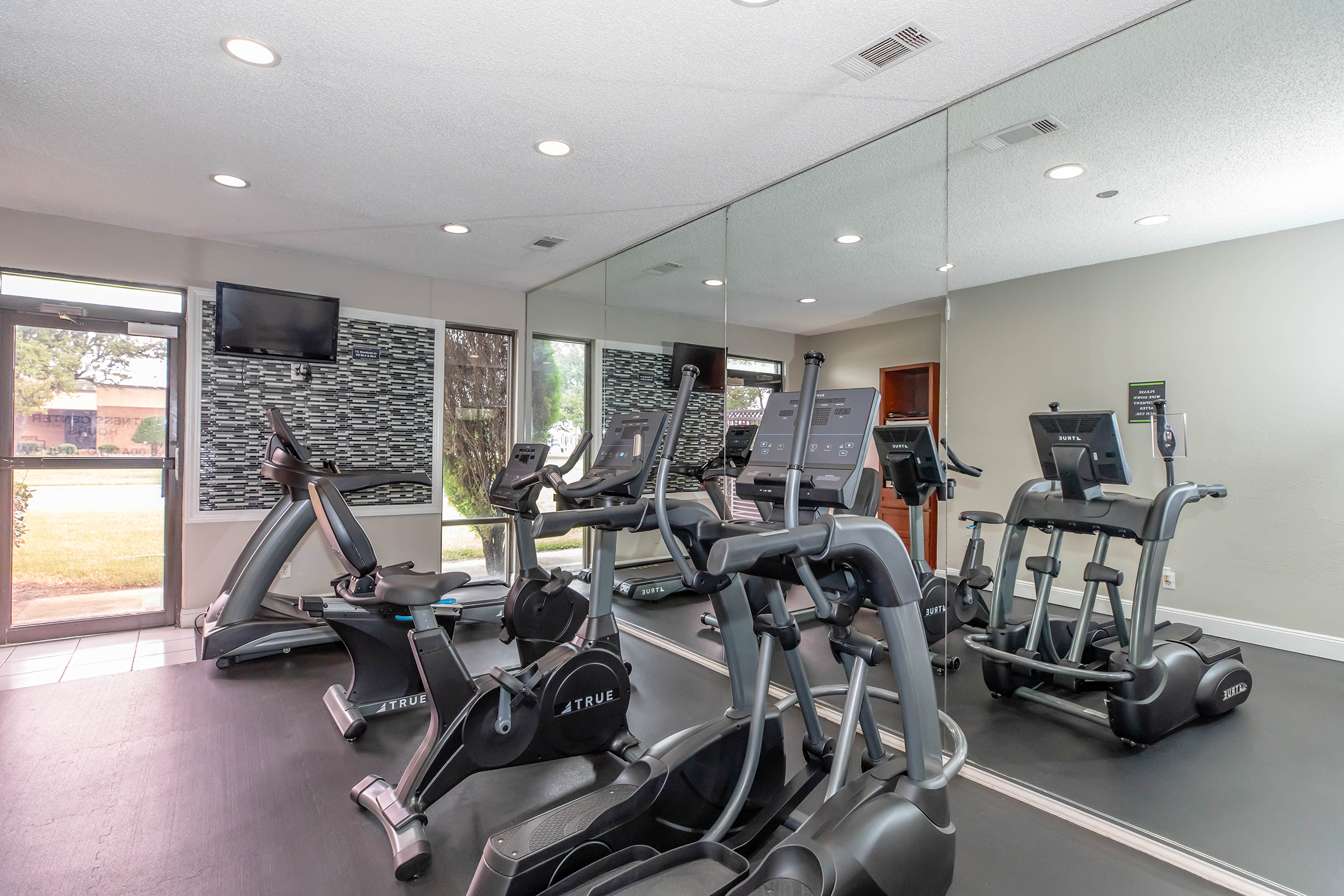 A well-lit gym featuring various exercise equipment, including treadmills and elliptical machines, set on a dark floor. Large mirrors line one wall, reflecting the machines and outdoor natural light coming through windows. There is a television mounted on the wall for entertainment while exercising.