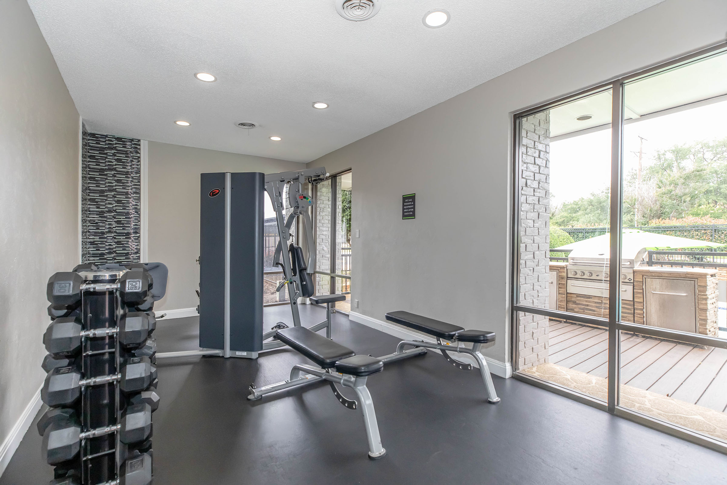 A modern gym interior featuring weight benches and a multi-exercise machine, with a rack of dumbbells. Large windows offer a view of an outdoor area with a barbecue grill. The space has neutral-colored walls and bright overhead lighting, creating an inviting atmosphere for fitness activities.